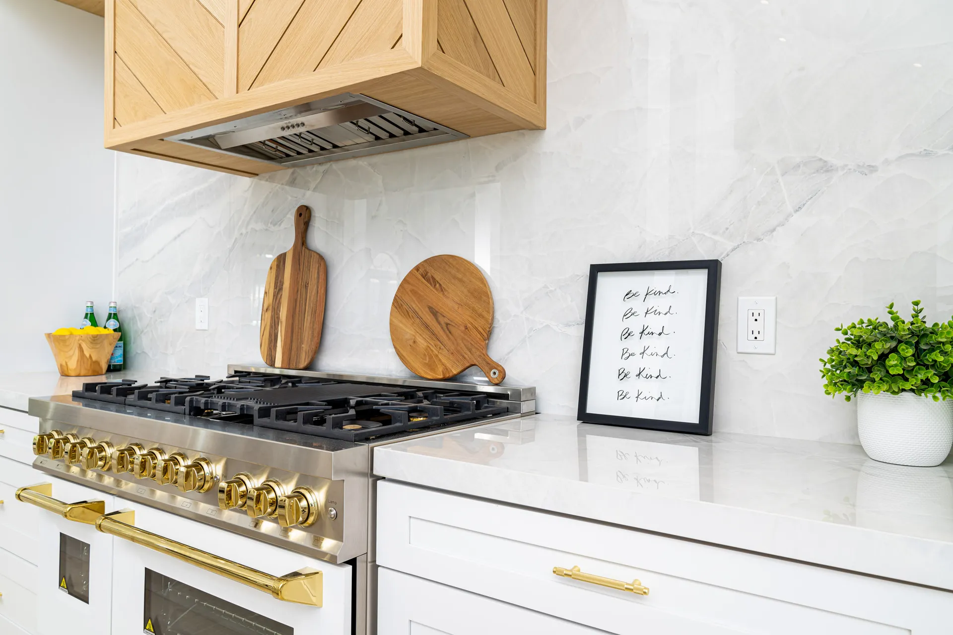 Kitchen countertop with a gas stove featuring gold accents, a wooden cutting board, a framed quote, and a plant. The countertop is made of white marble, and the cabinets have gold handles, creating a luxurious and elegant kitchen space.