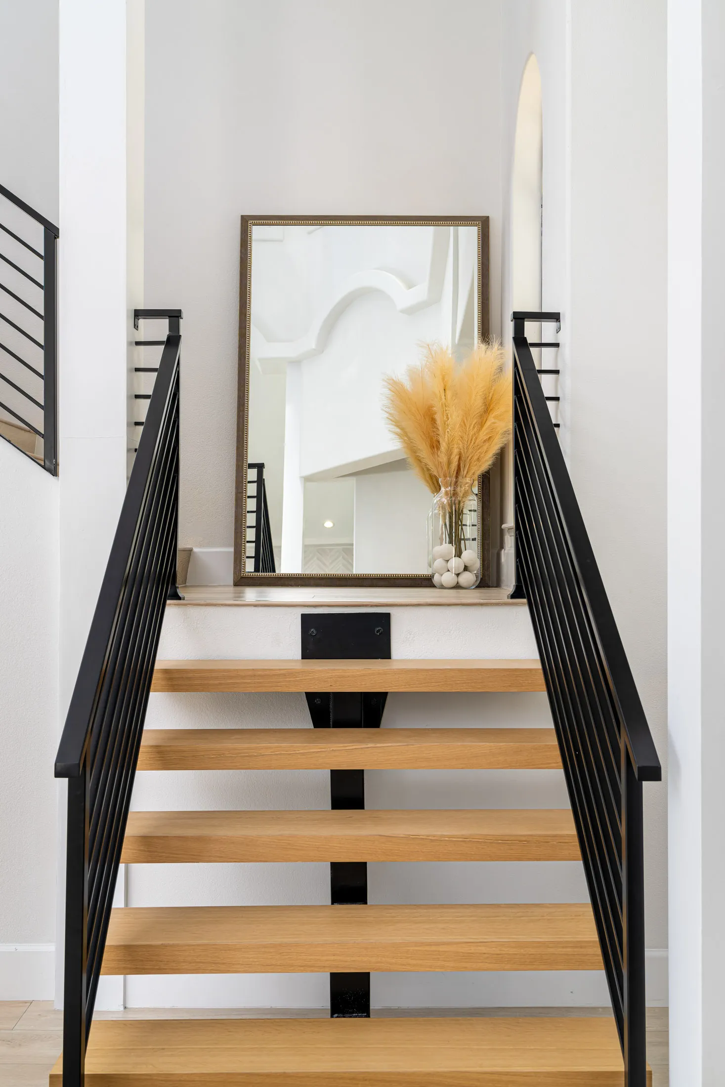Staircase leading up to a landing area with a large mirror and a vase with tall dried pampas grass. The stairs have wooden treads and black metal railings, and the landing area is brightly lit, creating an elegant and inviting entrance.