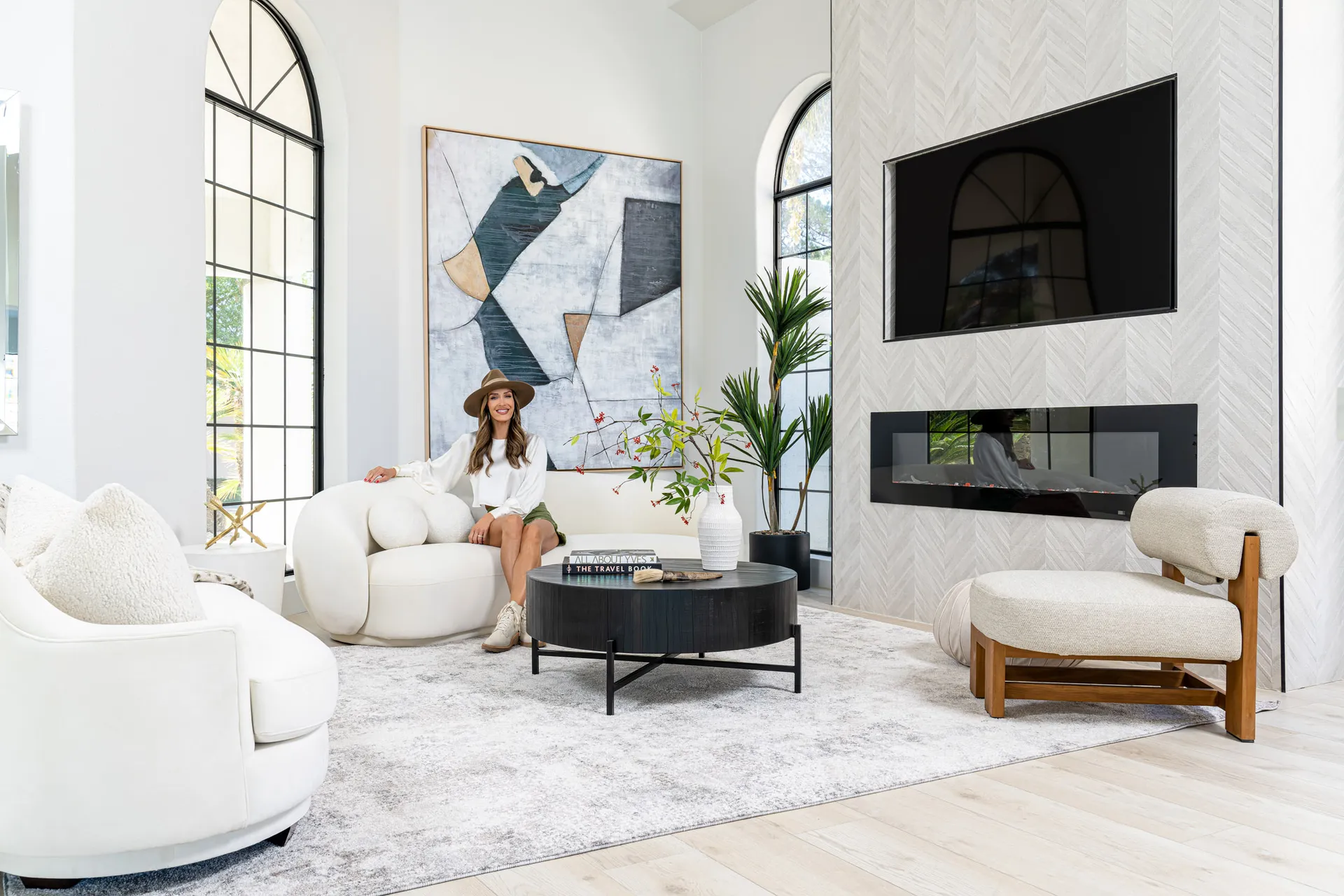 Person seated on a white curved sofa in a living area. The room features a black round coffee table, a large abstract painting, and a flat-screen TV mounted above a fireplace. Large windows provide natural light, and there are decorative items on the table, creating a stylish and inviting atmosphere.