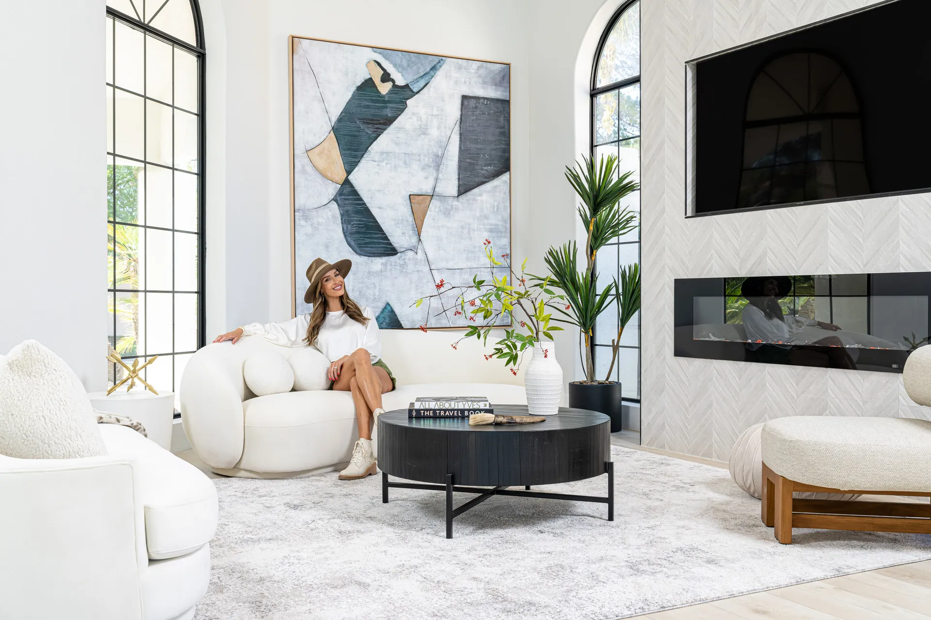 Person seated on a white curved sofa in a living area. The room features a black round coffee table, a large abstract painting, and a flat-screen TV mounted above a fireplace. Large windows provide natural light, and there are decorative items on the table, creating a stylish and inviting atmosphere.