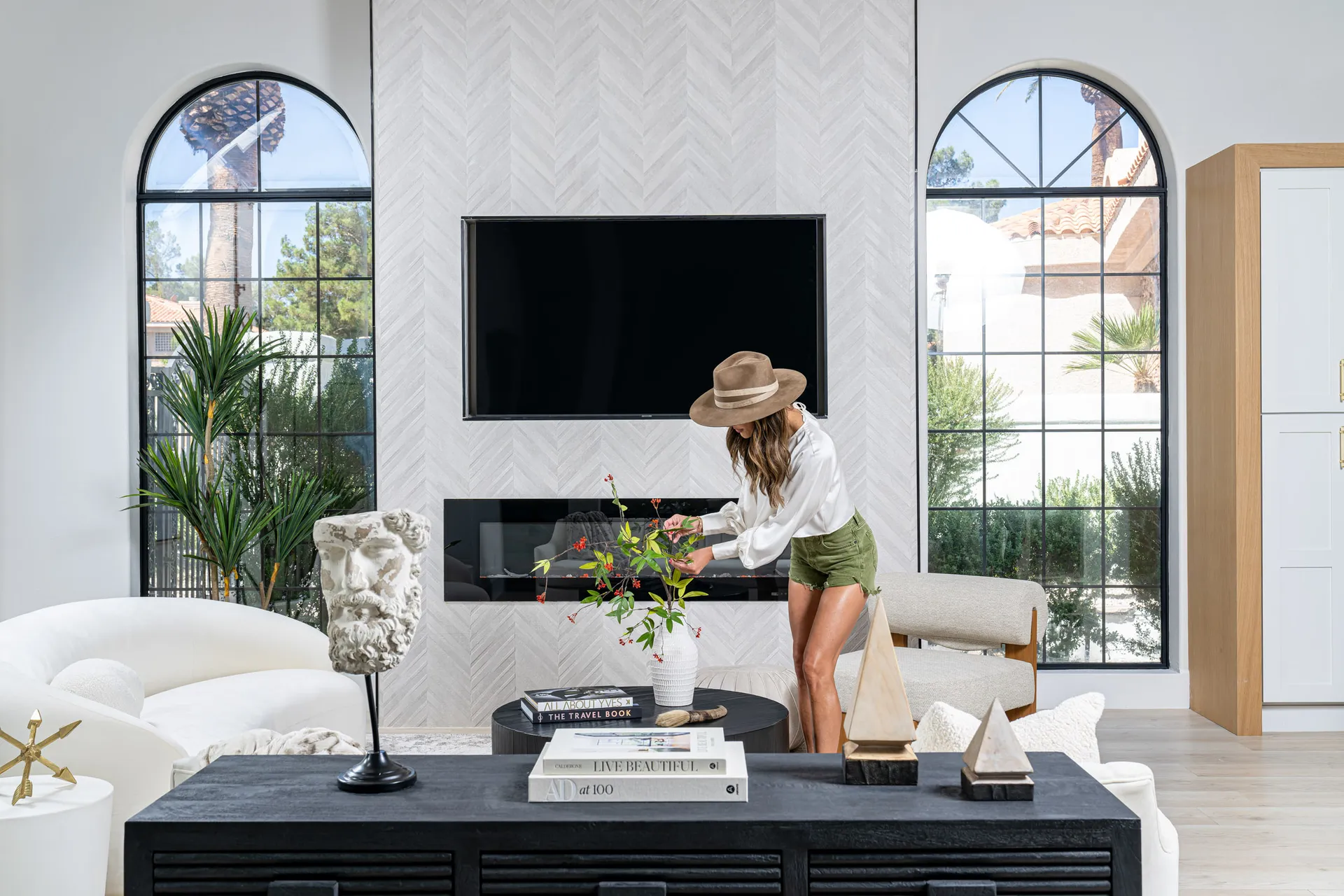 Person arranging flowers on a black coffee table in a living area. The room features a white curved sofa, a beige armchair, and a flat-screen TV mounted above a fireplace. Large windows provide natural light, and there are decorative items on the table, creating a stylish and inviting atmosphere.