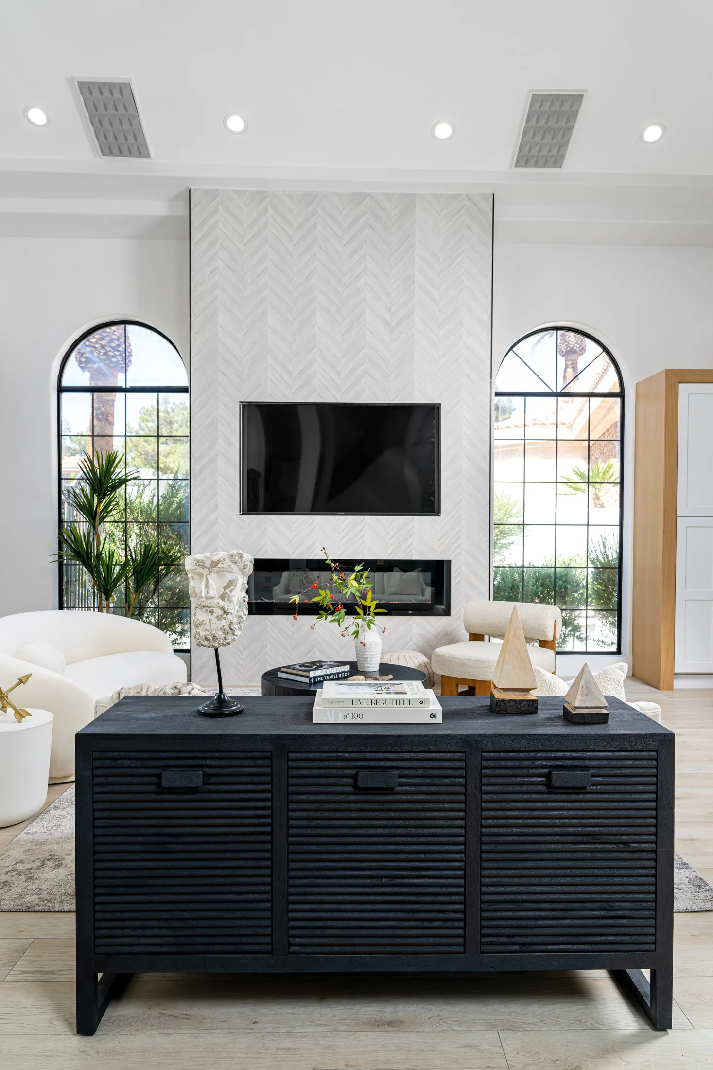Black console table with a textured surface, placed in front of a fireplace with a flat-screen TV mounted above it. The table has decorative items, including a vase with flowers and geometric wooden sculptures. The room has high ceilings and large windows, creating a bright and modern atmosphere.