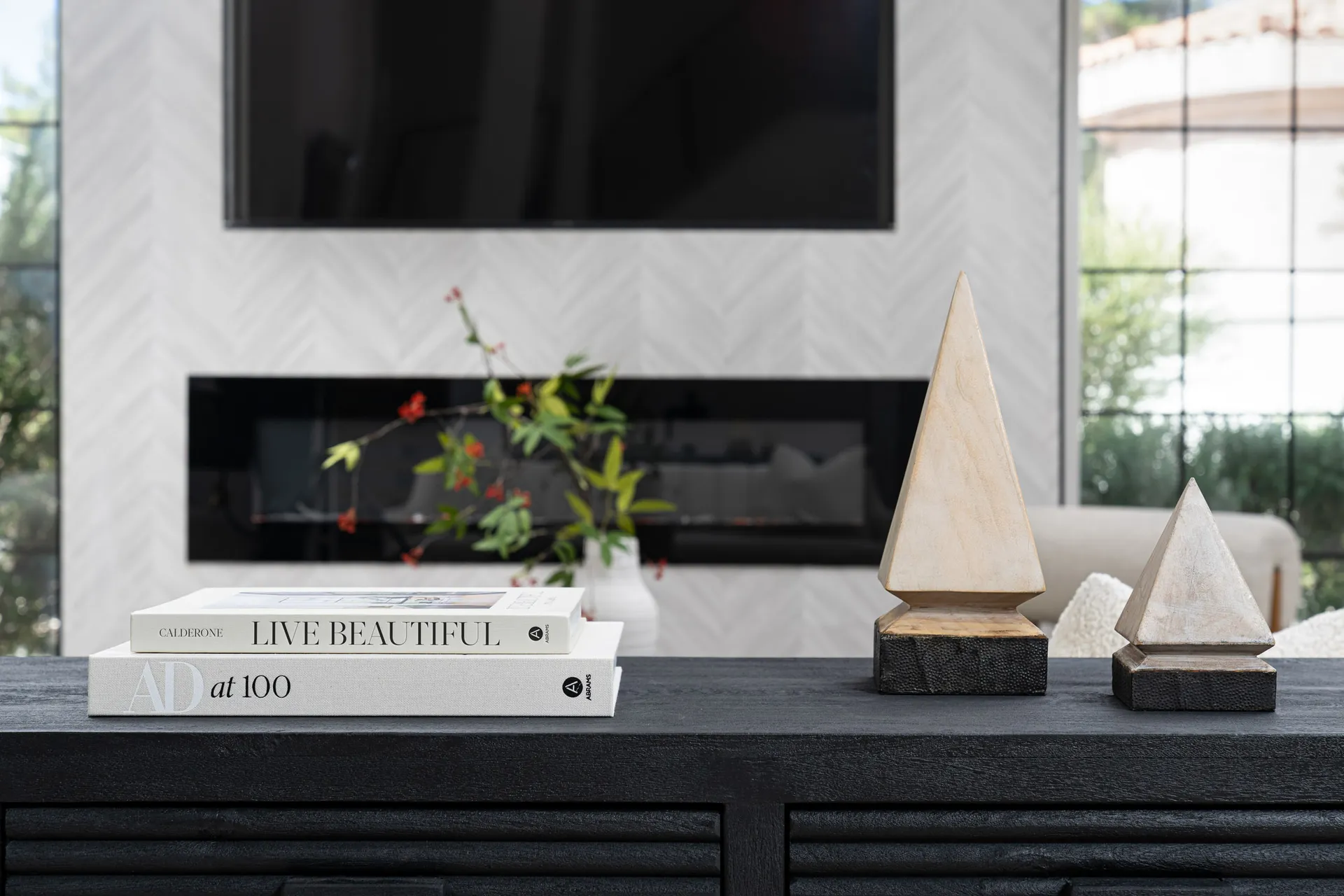 Close-up of a black console table with a stack of books and a geometric wooden sculpture. In the background, there is a fireplace with a flat-screen TV mounted above it, adding a modern touch to the space.