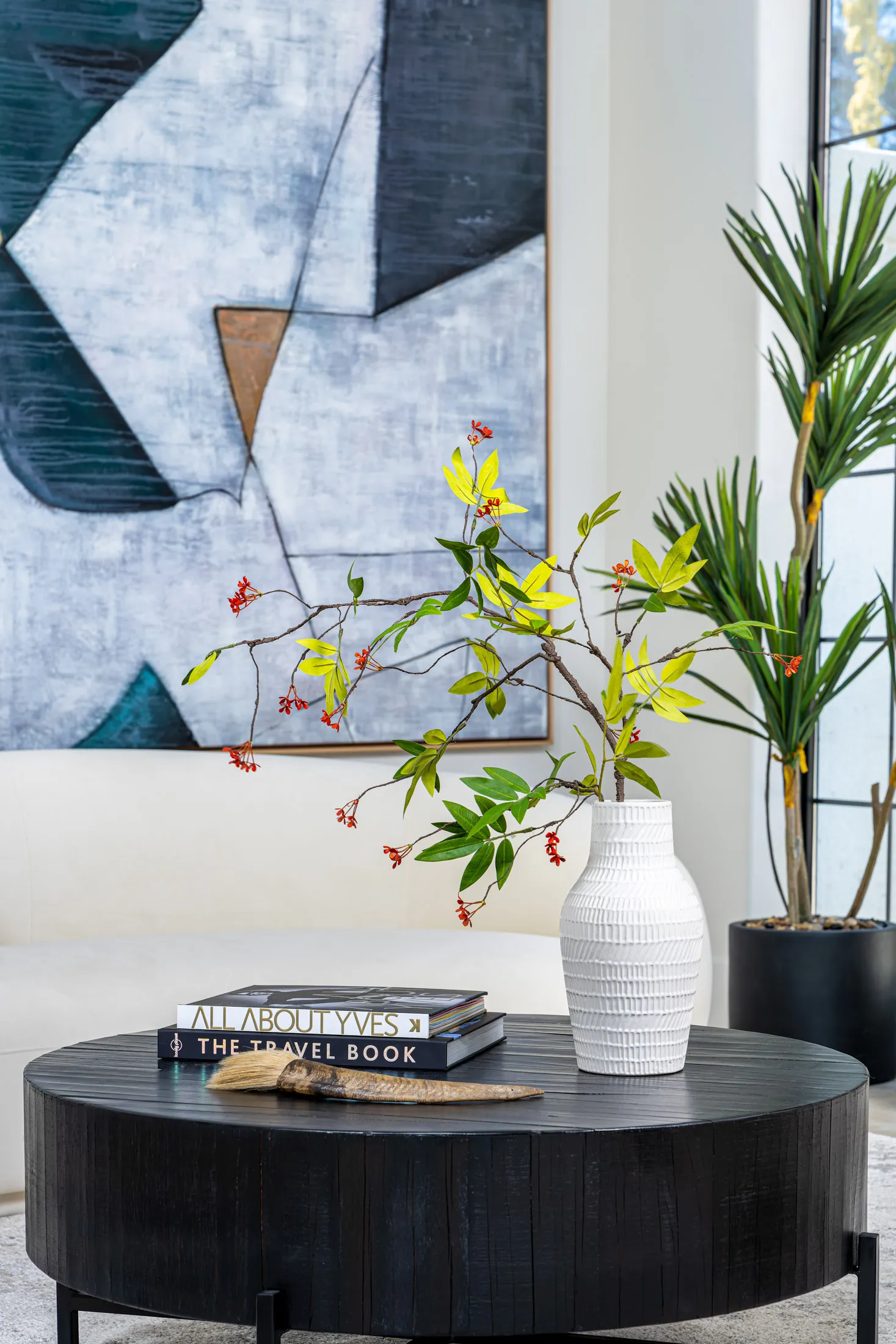 Close-up of a black round coffee table with a stack of books and a white vase containing a floral arrangement. In the background, there's a large abstract painting, adding an artistic touch to the setting.