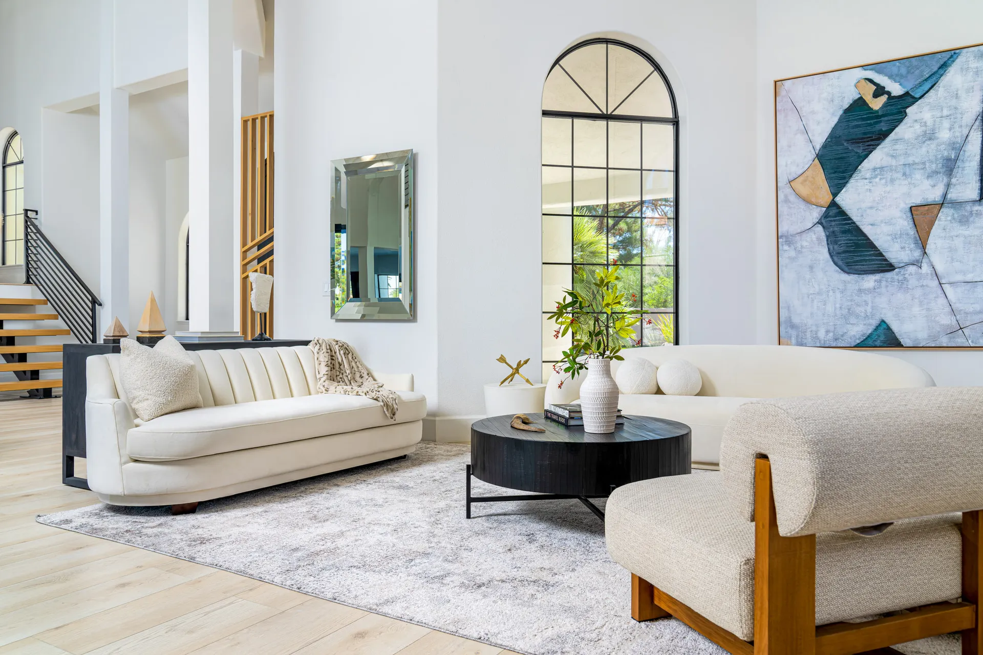 Living area with a white sofa and a beige armchair arranged around a round black coffee table. There's a large abstract painting on the wall, and a tall mirror is placed near the window. The room has high ceilings and large windows that provide natural light, creating a bright and spacious atmosphere.