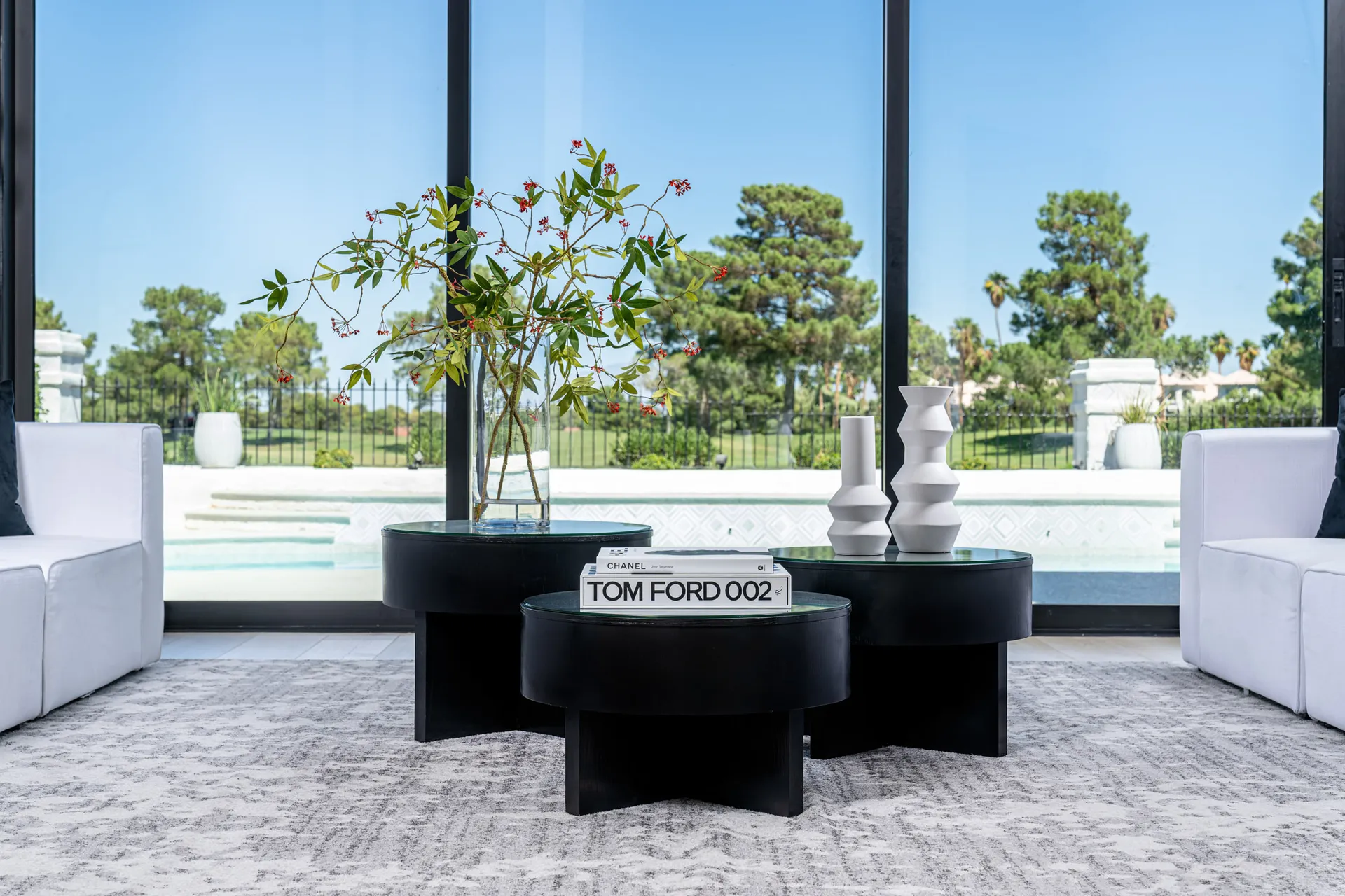 Seating area with black coffee tables arranged on a gray rug. On one of the tables, there's a stack of books with a vase containing a floral arrangement. Large windows provide a view of an outdoor space with a swimming pool, creating a serene and elegant setting.