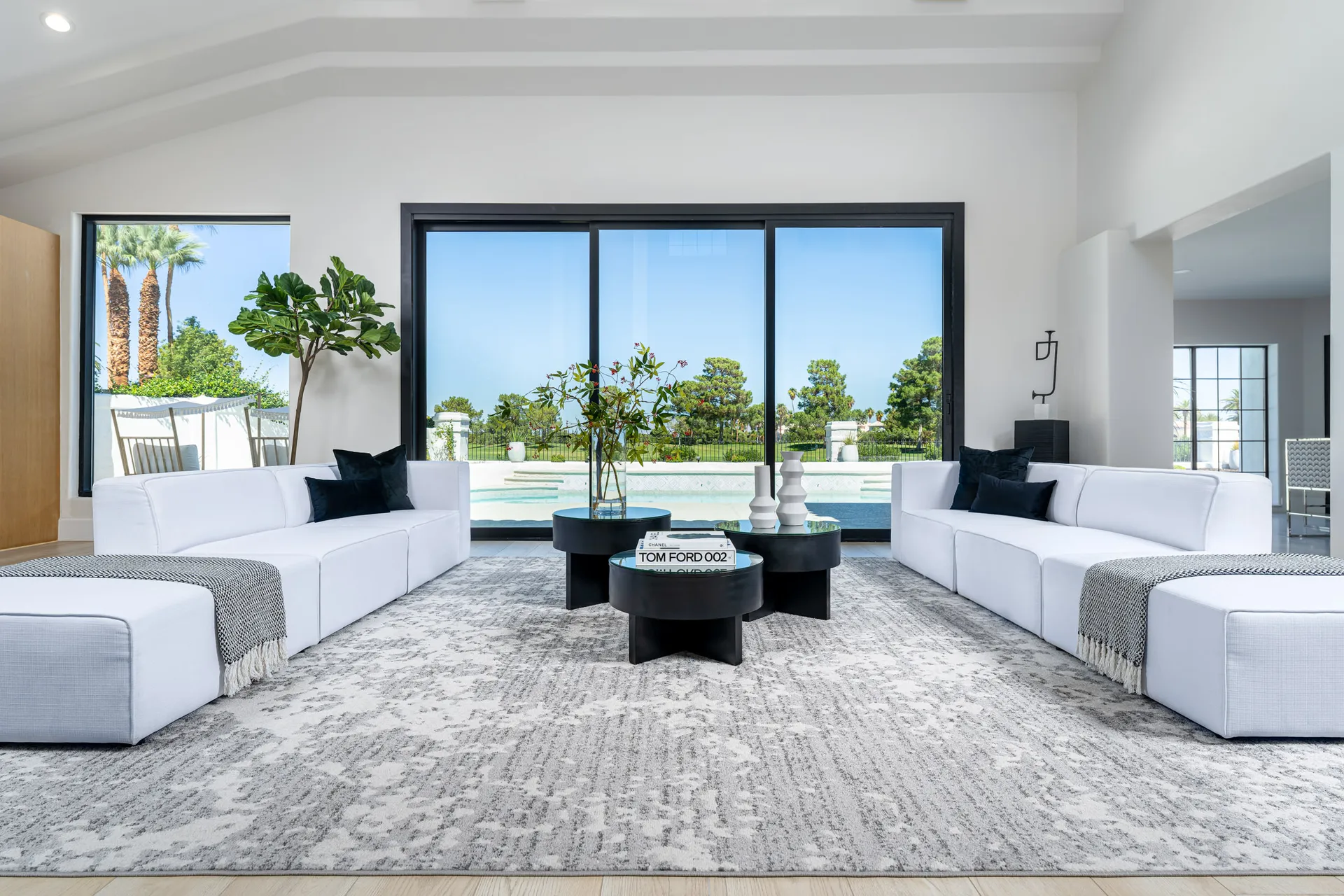 Large open space with white sofas arranged around black coffee tables on a gray rug. The area has high ceilings, large windows, and a chandelier. There's also a dining area and a kitchen visible in the background, creating a spacious and elegant atmosphere.