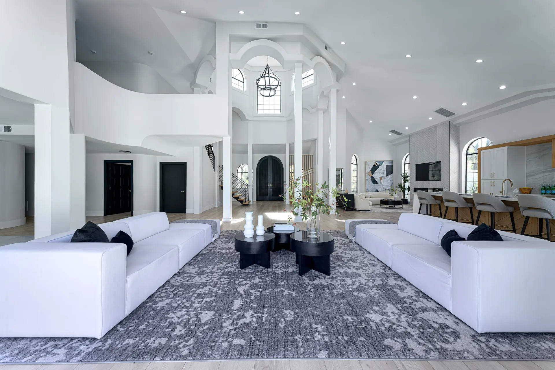 Large open space featuring white sofas arranged around black coffee tables, placed on a gray rug. The area has high ceilings, large windows, and a chandelier, with a dining area and kitchen visible in the background. The design is elegant and spacious, creating an inviting atmosphere.