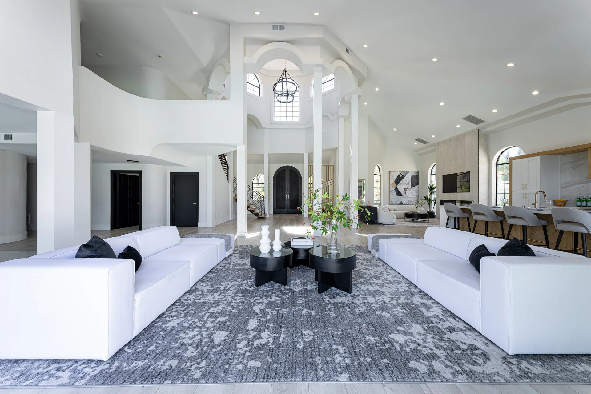 Large open space with white sofas and a gray rug, arranged around a black coffee table. The area features high ceilings, large windows, and a chandelier. There's also a dining area and a kitchen visible in the background, creating a spacious and elegant atmosphere.