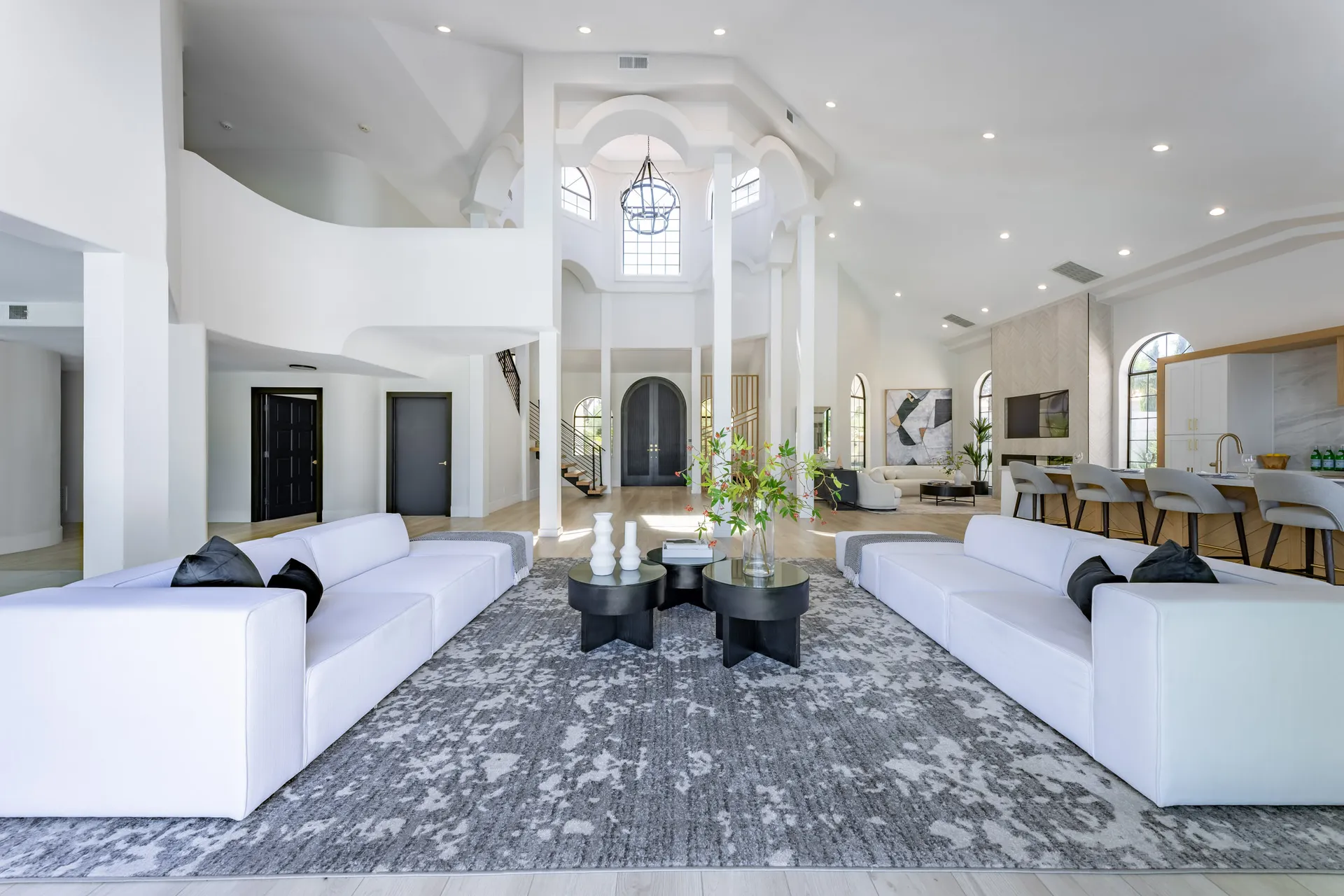 Spacious living area with white sofas arranged around a black coffee table. The room features high ceilings, large windows, and a chandelier. There's also a dining area and a kitchen visible in the background, creating an open and inviting space.