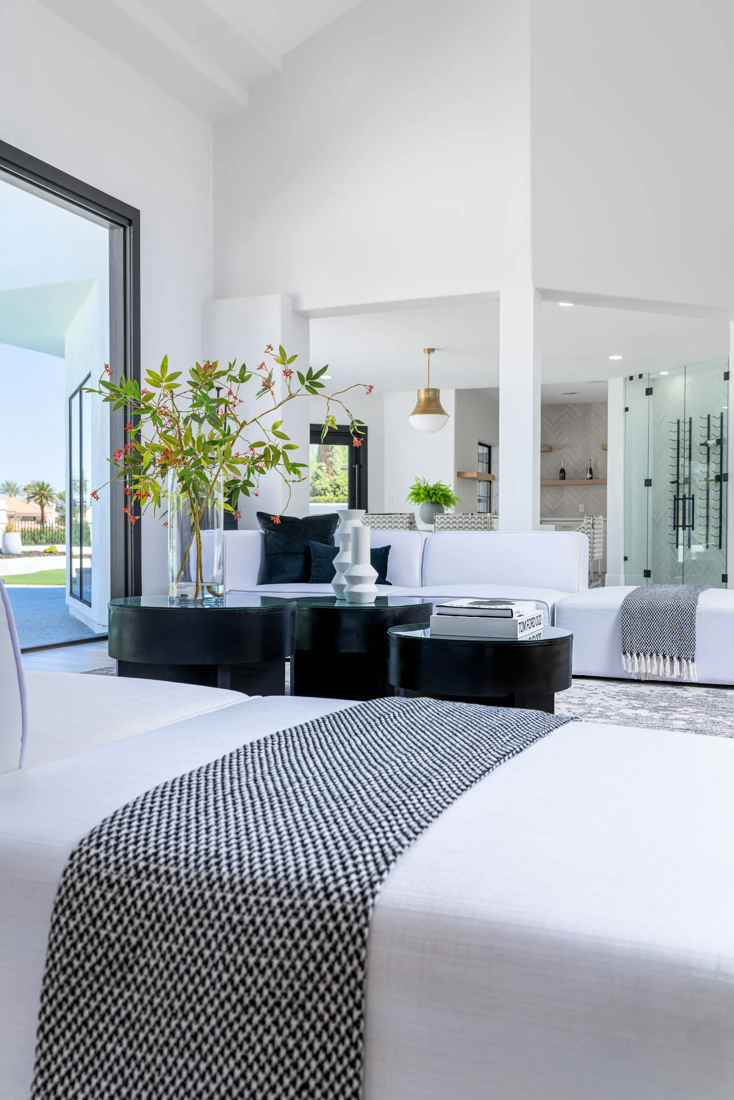 Bedroom with a view of a seating area. There's a bed with white bedding and a gray throw, and in the background, there's a seating area with white sofas, a black coffee table, and a vase with flowers. Large windows provide natural light, creating a bright and airy atmosphere.
