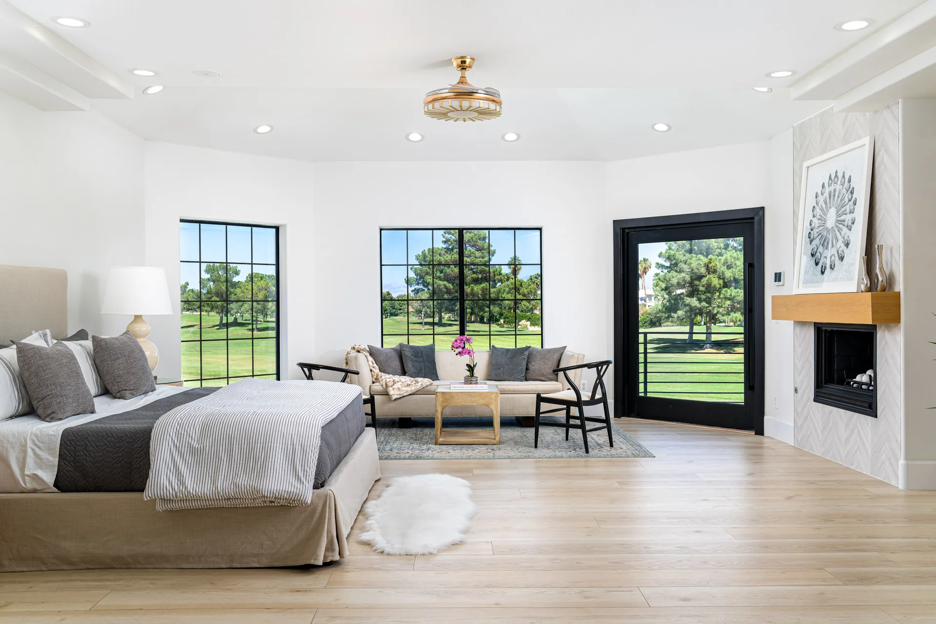 Spacious living area with a modern design. There's a beige sectional sofa with gray and white throw pillows, a glass coffee table, and a fireplace with a herringbone-patterned wall. Large windows provide natural light, and there's a chandelier hanging from the ceiling, creating a comfortable and inviting space.