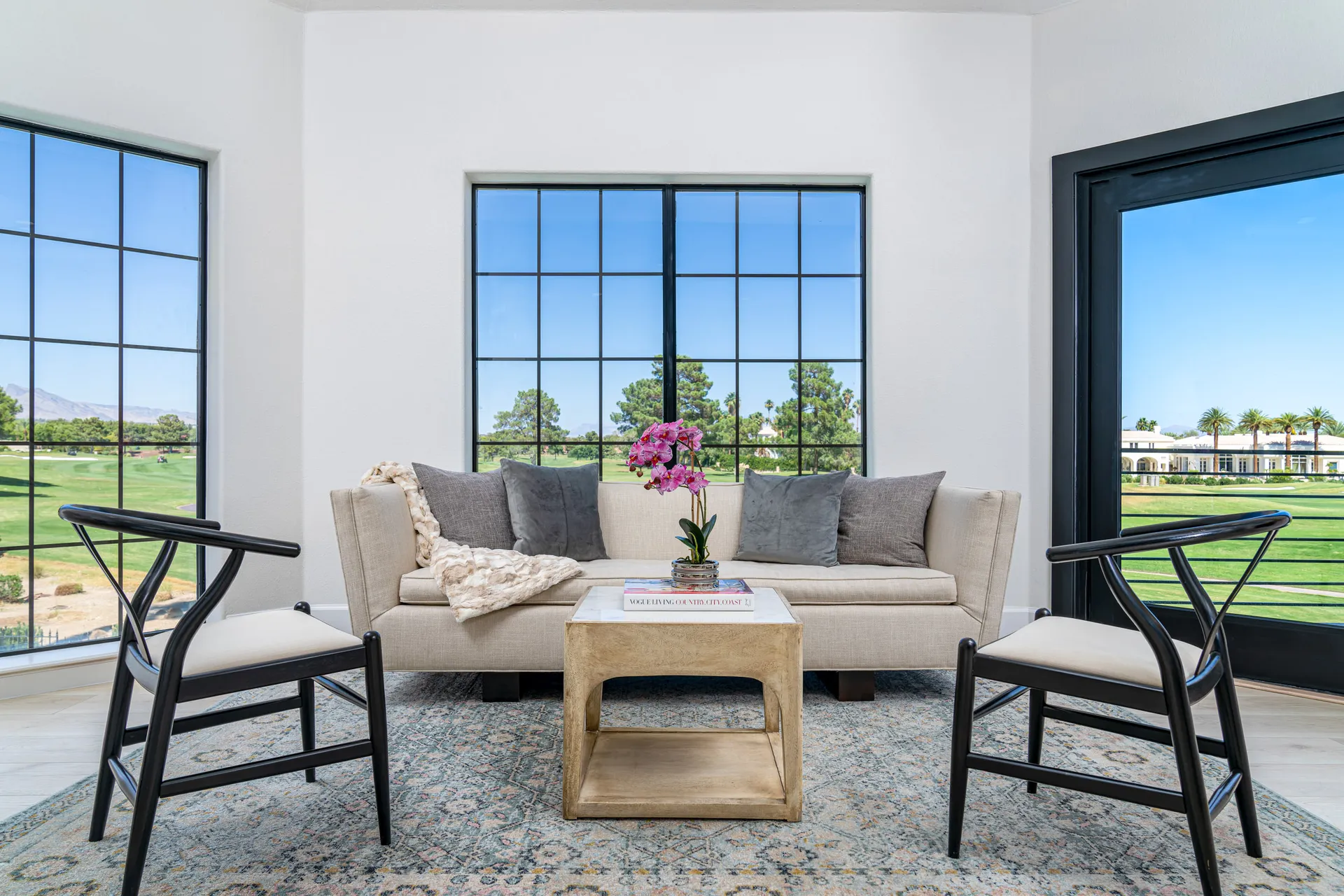 A neutral couch with an orchid on a square coffee table, flanked by black chairs and large windows overlooking a golf course.