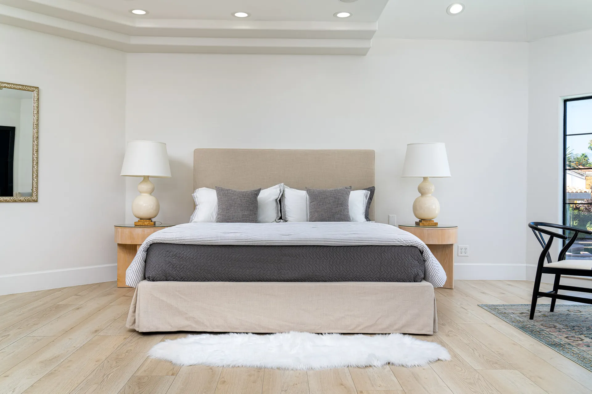 Bedroom with a large bed featuring a beige headboard and white bedding, complemented by gray throw pillows. The room has wooden flooring, nightstands with lamps, and a large window that provides natural light. The design is modern and comfortable, creating a relaxing atmosphere.
