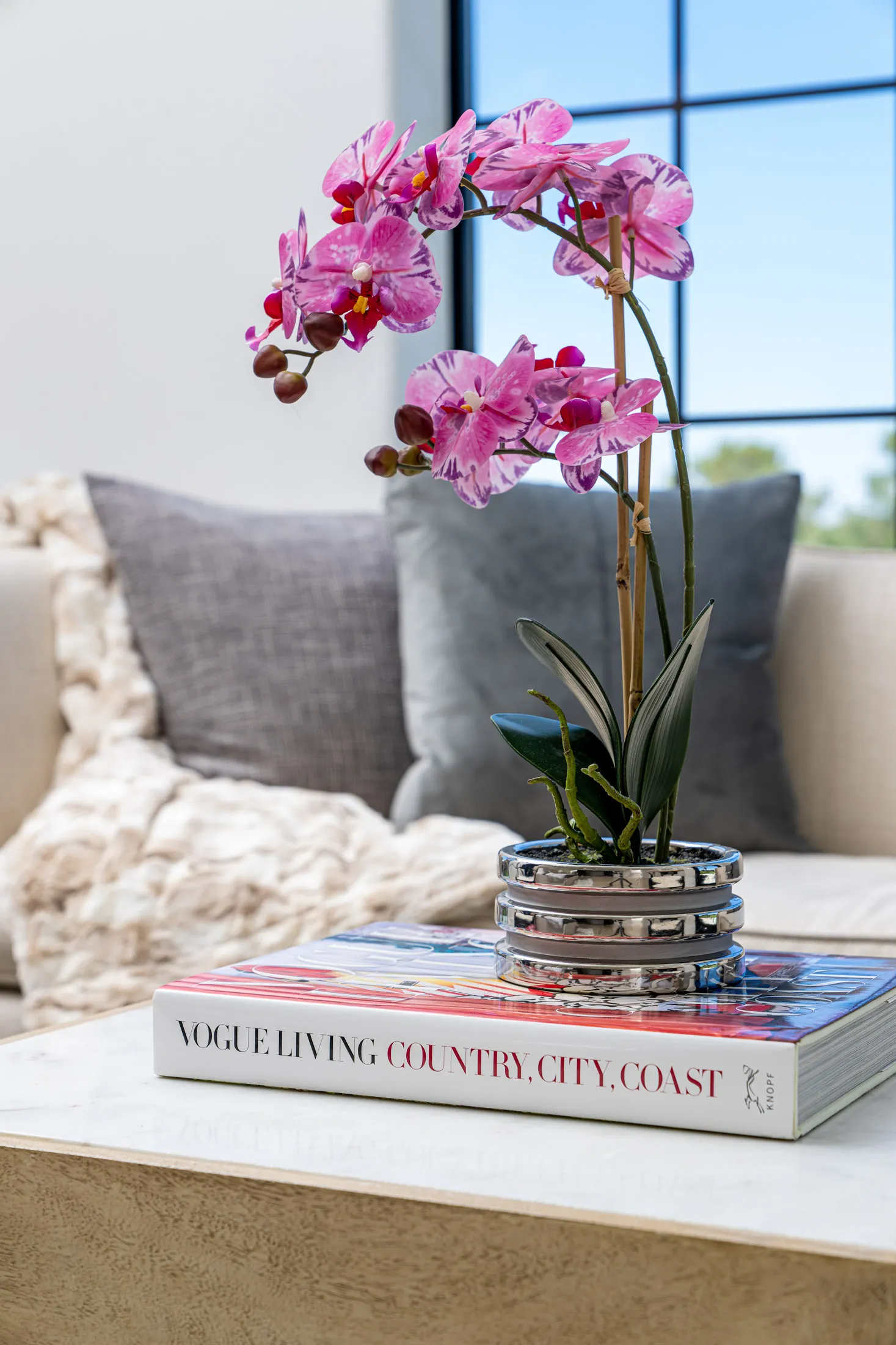 Close-up of a potted orchid plant placed on top of a stack of books on a marble table. The background features a sofa with cushions and a blanket, and a large window provides natural light. The arrangement adds a touch of elegance and nature to the space.