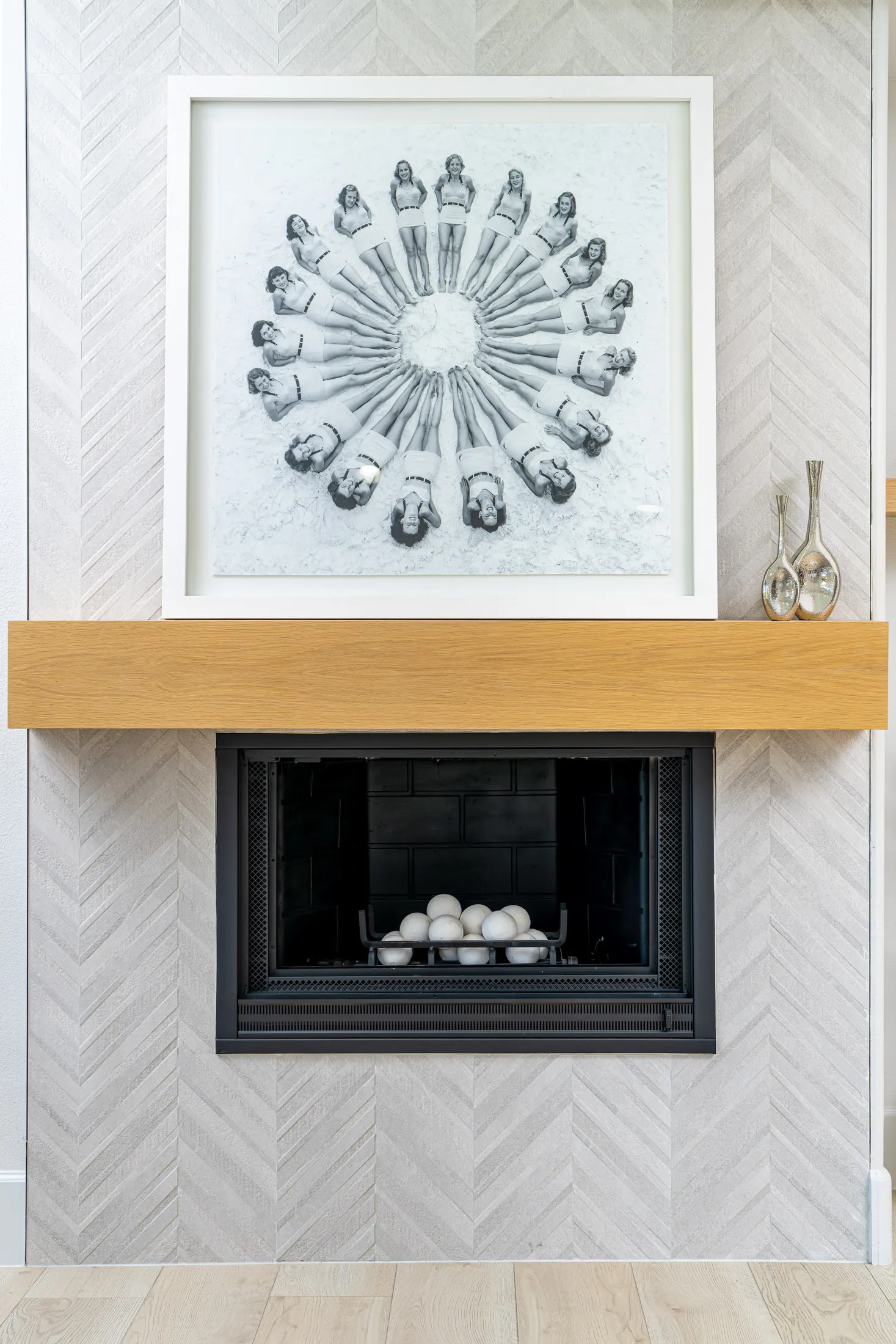 Fireplace with a wooden mantel and a modern art piece hanging above it. The fireplace is set against a light-colored herringbone-patterned wall, and there are decorative vases on the mantel. The design is contemporary and adds warmth to the space.