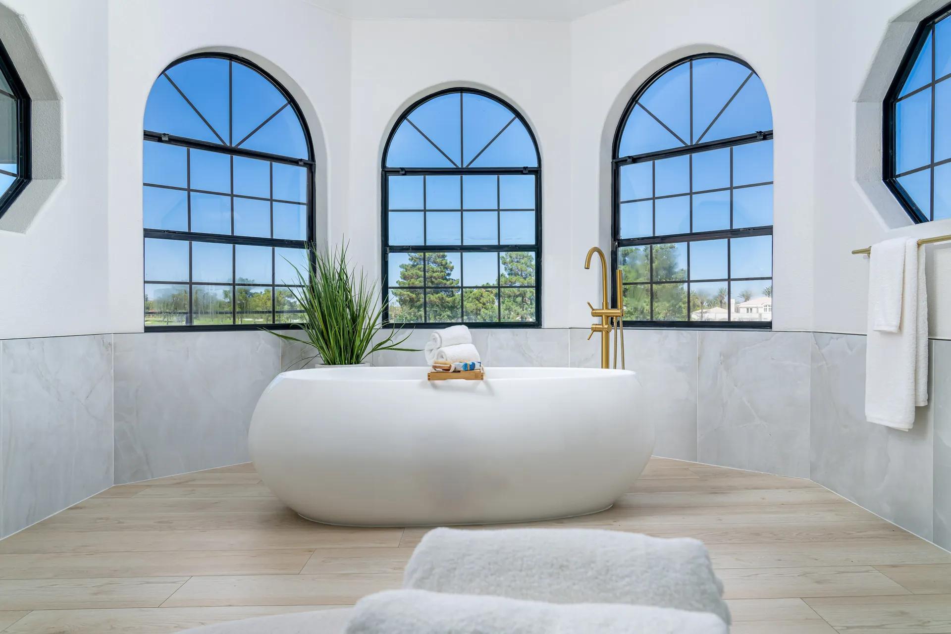 Luxurious bathroom with a freestanding white bathtub, surrounded by large arched windows. The floor is wooden, and there's a potted plant beside the tub, adding a touch of greenery. The space is filled with natural light, creating a serene and relaxing atmosphere.