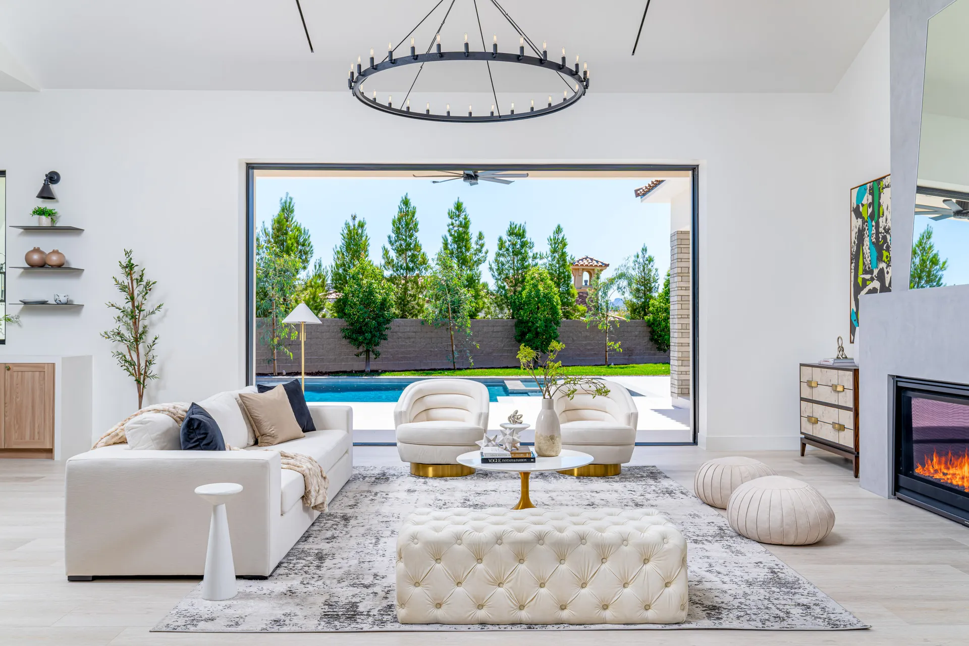 A living rooA living room with a large window overlooking a pool and greenery, white sofas, a tufted ottoman, and a fireplace, creating a luxurious and inviting atmosphere. The design features clean lines, a neutral color palette, and an open layout.m with a large window overlooking a pool and greenery, white sofas, a round coffee table, and a tufted ottoman. The design features a modern and elegant aesthetic, with a serene view of the outdoors.