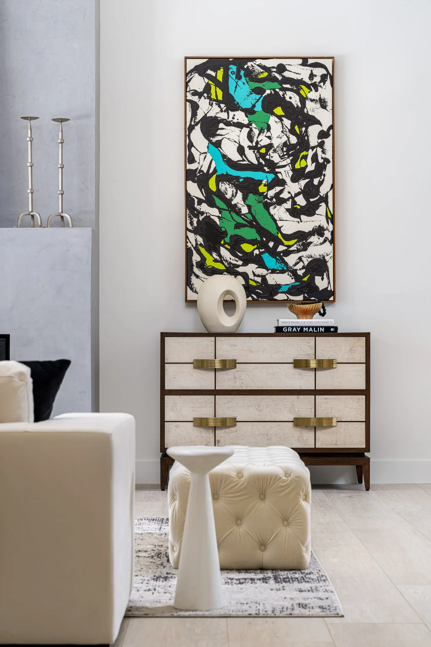 A light-toned dresser with gold hardware stands beneath a large abstract painting in vibrant colors. A neutral ottoman and candlesticks are nearby.