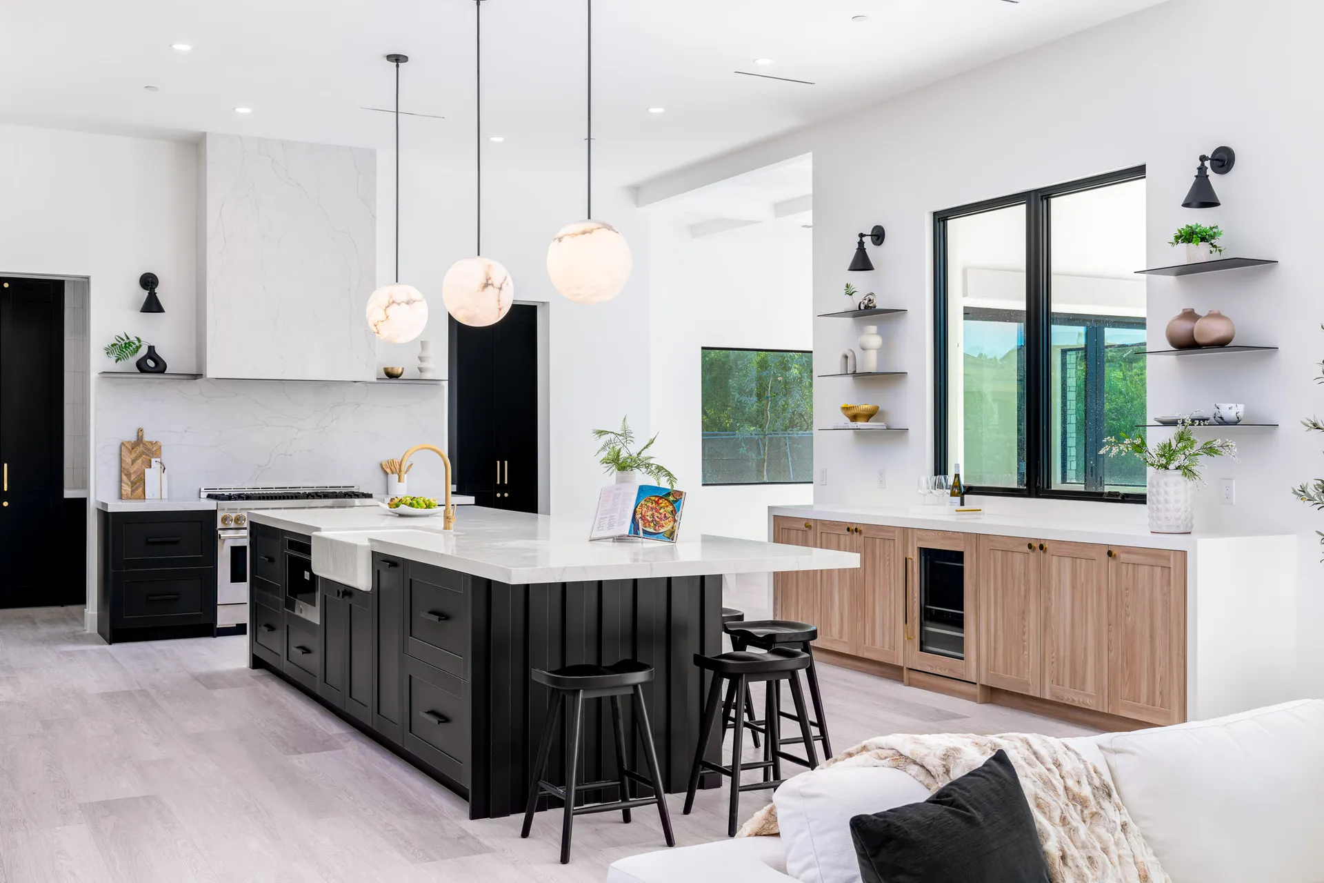 A kitchen with a black island and white marble countertops, accented by brass hardware and three round pendant lights overhead. Light wood cabinetry and open shelves add warmth and balance.