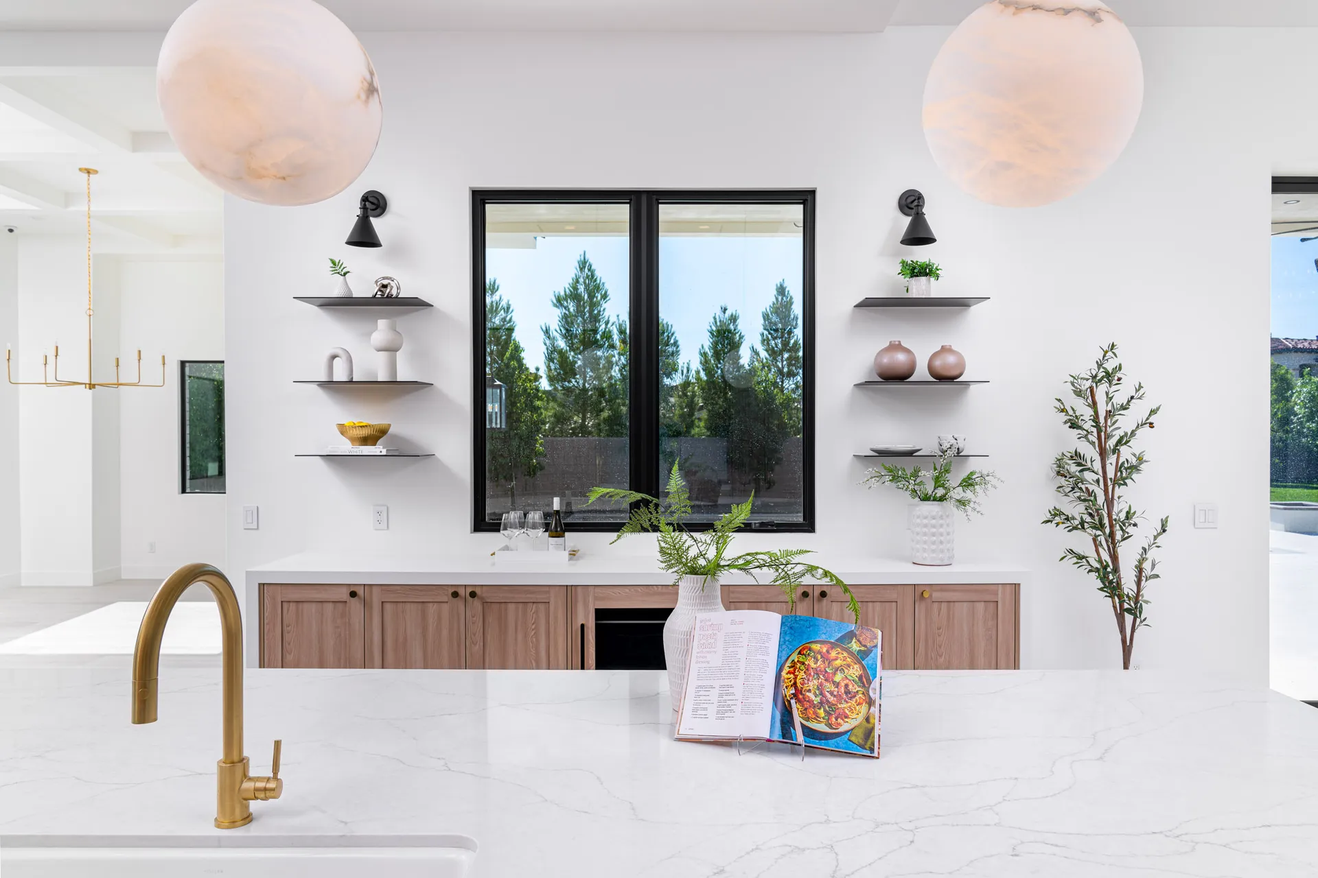 A kitchen island with a gold faucet and white marble countertop. Floating shelves around a central window display decorative vases and modern accents.