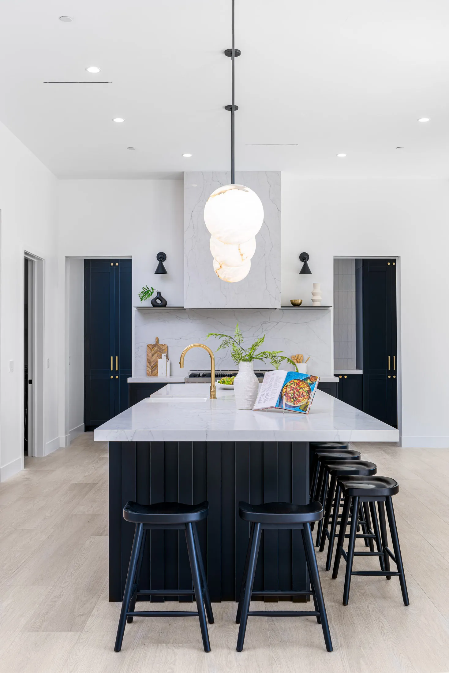 A kitchen island with black vertical paneling, topped by a white marble counter, with matching black stools lined up. Three round pendant lights hang above, and gold hardware accents the faucet and cabinetry.