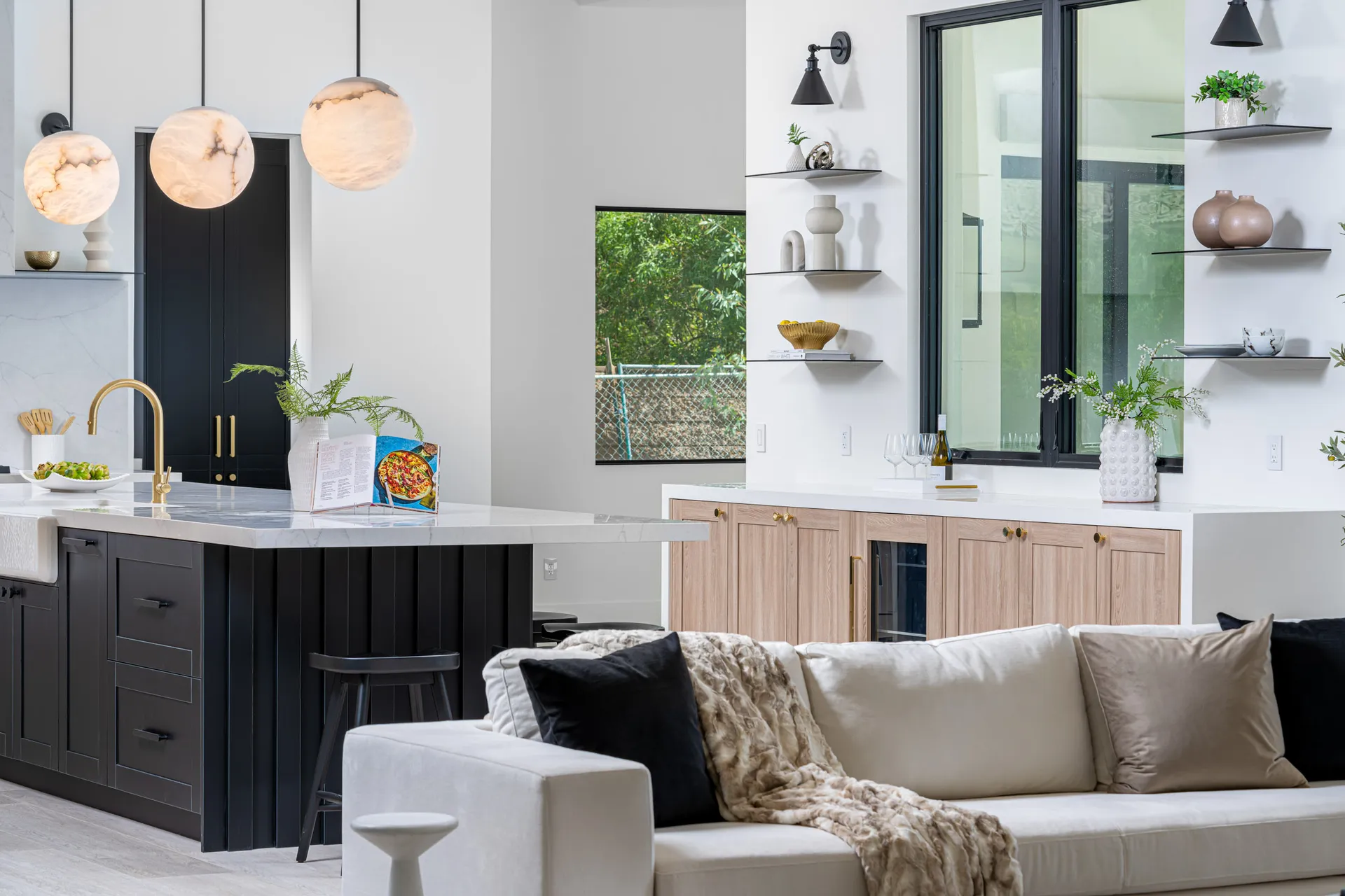 A kitchen from a Luxurious Comfort Home Staging project, featuring black lower cabinets, a white marble peninsula with a gold faucet, and light wood cabinetry with floating shelves.
