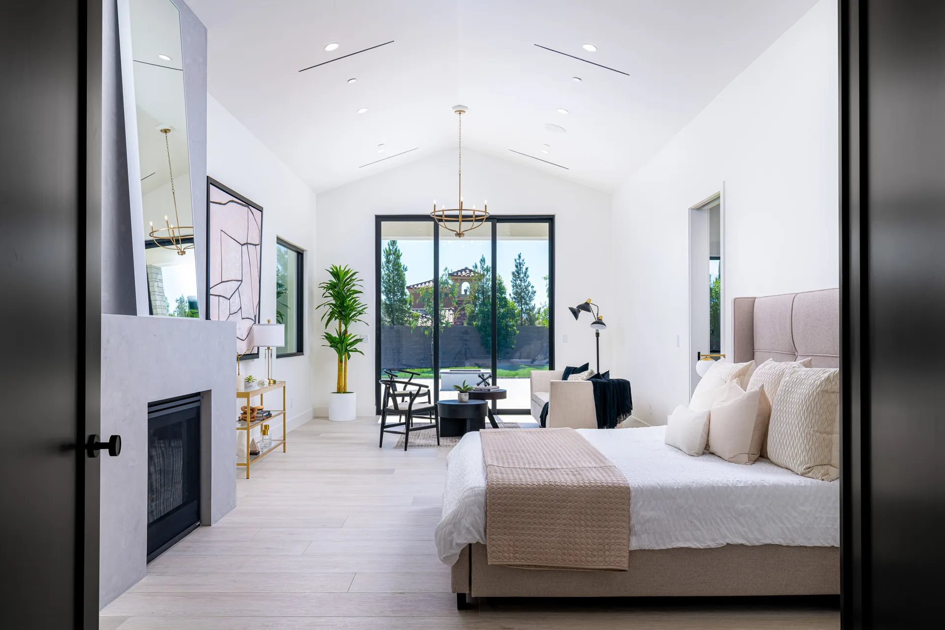 A bright bedroom with a vaulted ceiling, neutral bedding, and a modern fireplace. Large sliding doors open to a view of greenery.