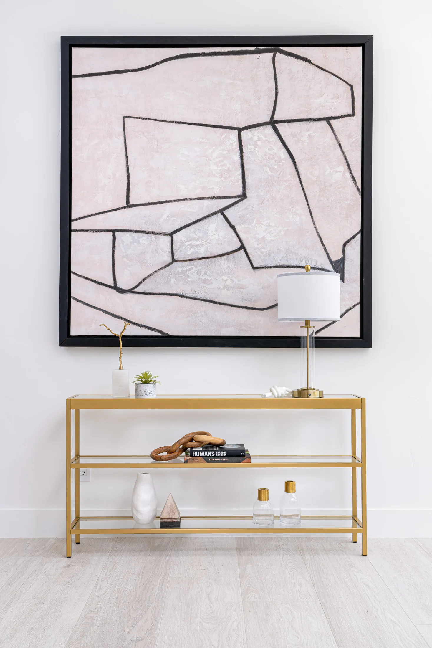 A gold console table displaying a small lamp, a potted plant, and a wooden chain accent on stacked books. A large abstract painting with black lines hangs above it.