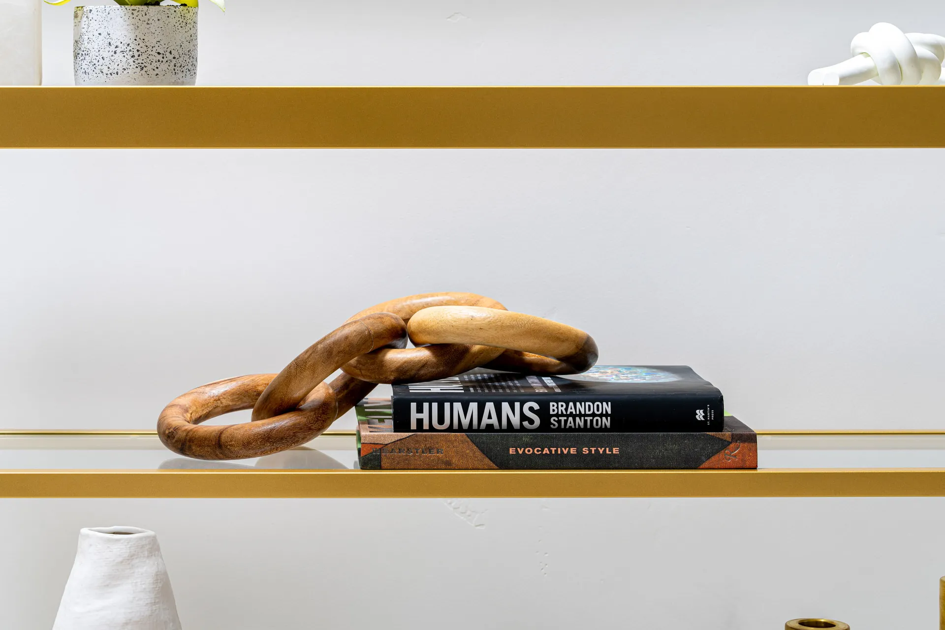A gold-framed glass shelf displaying two stacked books titled ‘Humans’ and ‘Evocative Style,’ topped with a wooden chain link accent. The shelf sits against a neutral wall.