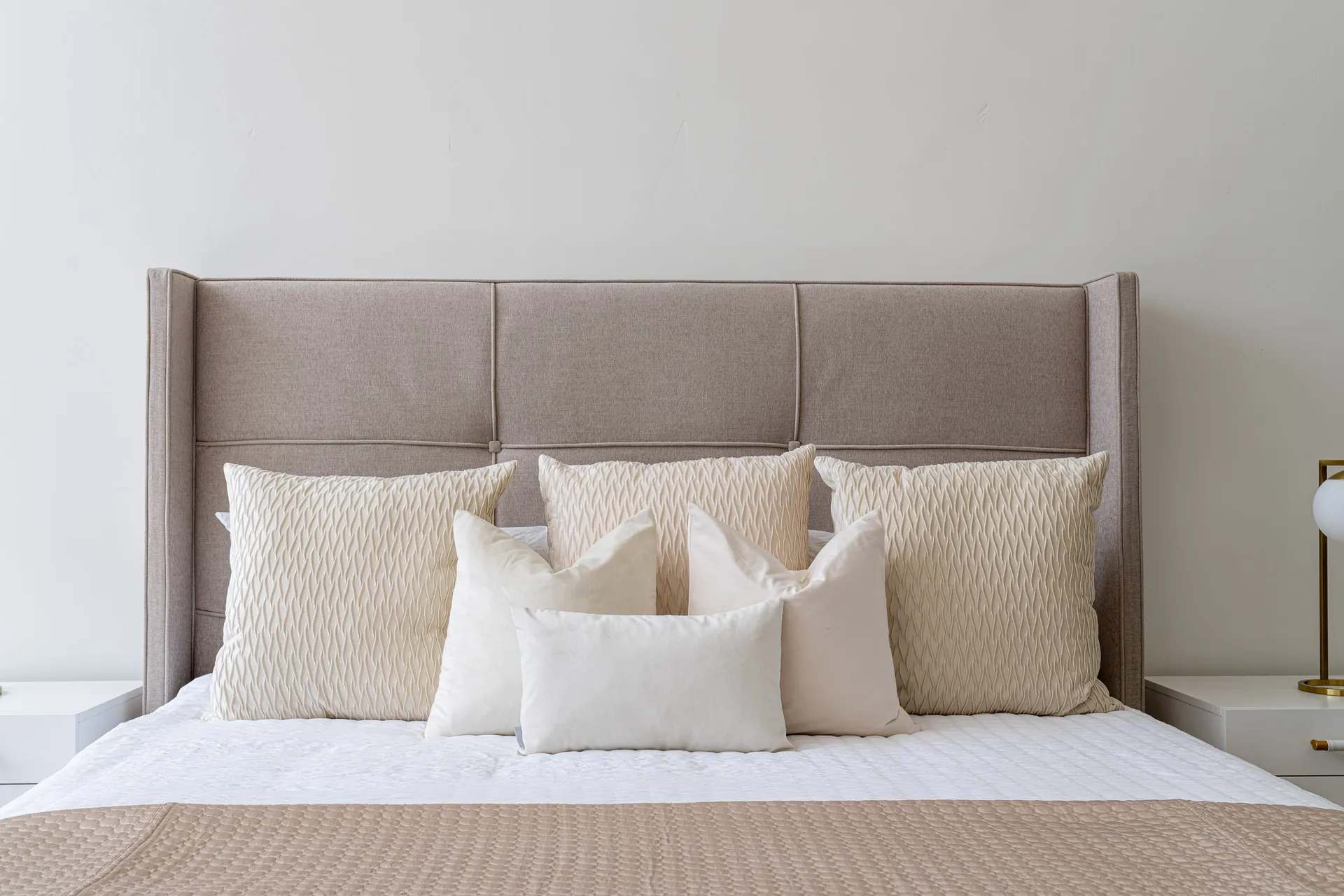 A tall wingback headboard in a soft beige fabric, layered with cream and ivory pillows. The bedding features subtle textures in coordinating neutral tones.