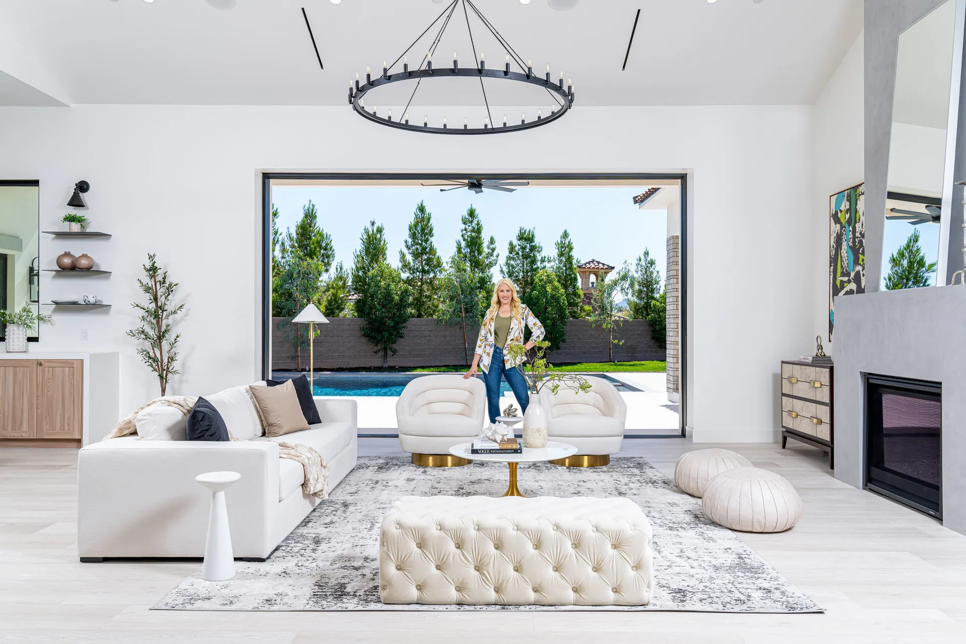 Kelly Long, Lead Interior Designer at Stately, standing in a modern living room with a white sofa and a view of a pool and garden. The room is stylishly designed with a fireplace, chandelier, and rug.