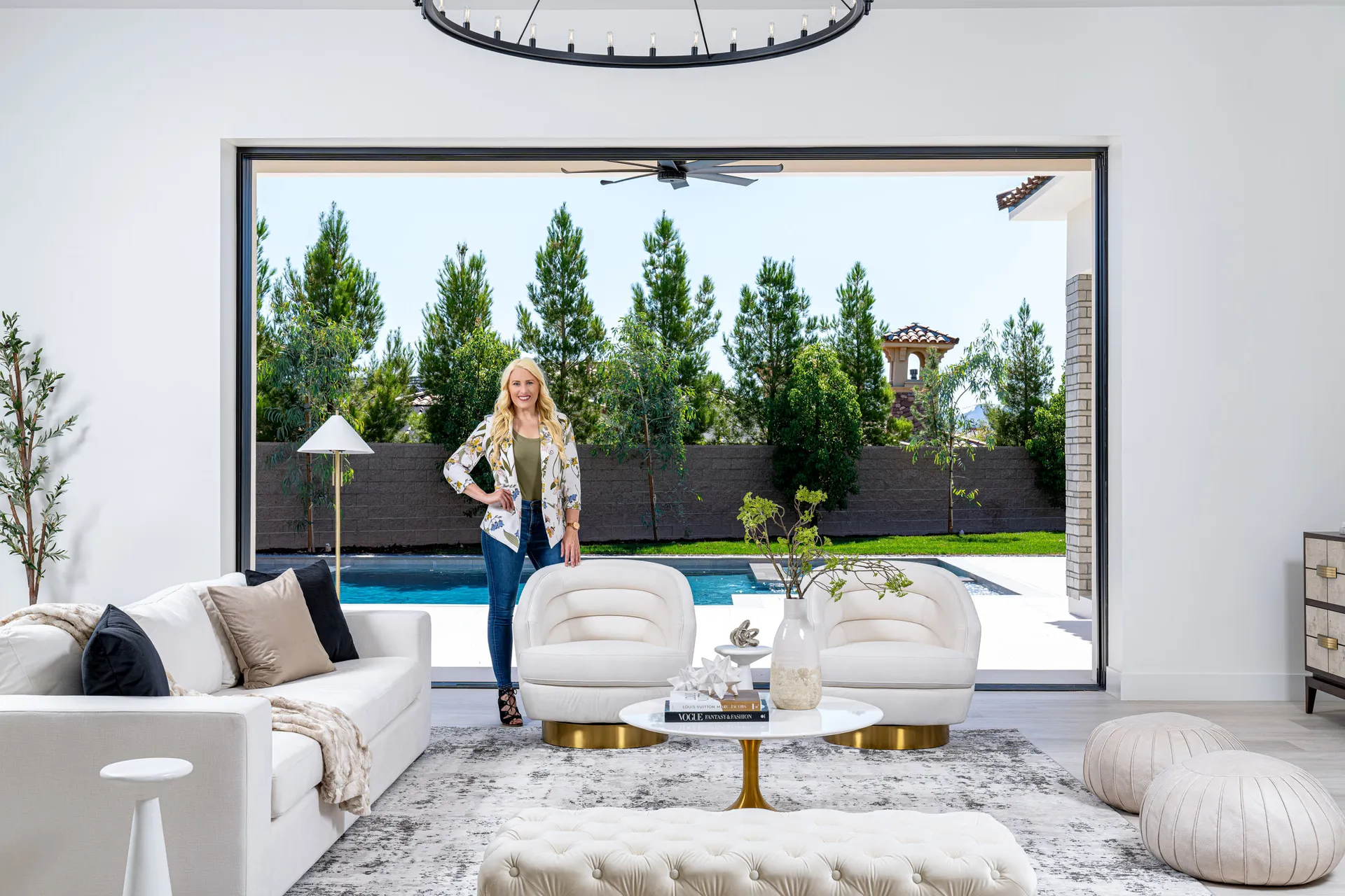 Kelly Long, Lead Interior Designer at Stately, standing in a modern living room with a white sofa and a view of a pool and garden. The room is stylishly designed with a fireplace, chandelier, and rug.