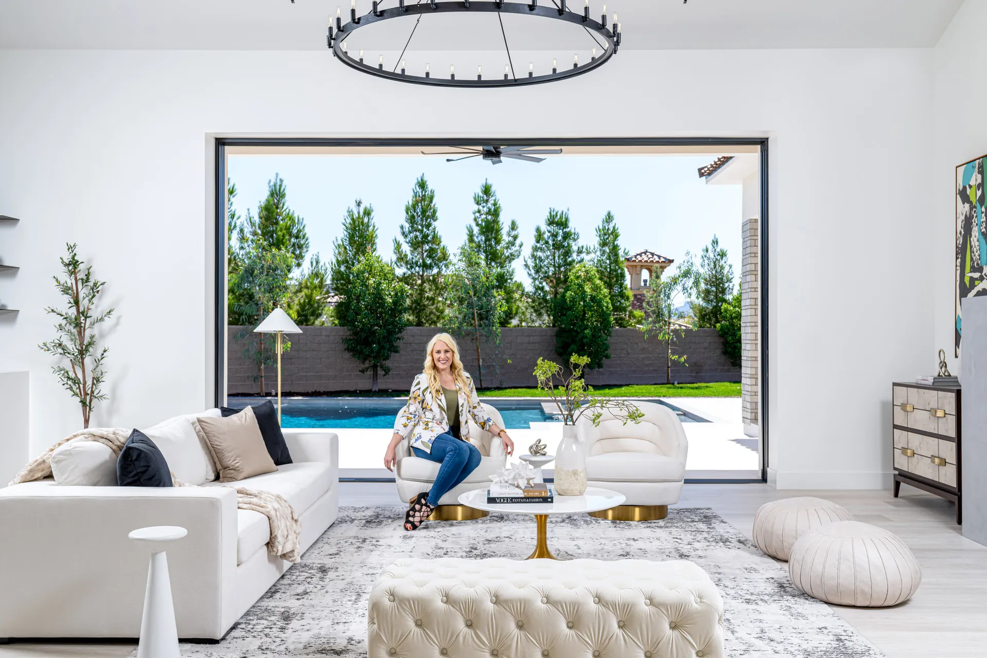 Kelly Long, Lead Interior Designer at Stately, sitting on a white sofa in a modern living room with a view of a pool and garden. The room is stylishly designed with a fireplace, chandelier, and rug.
