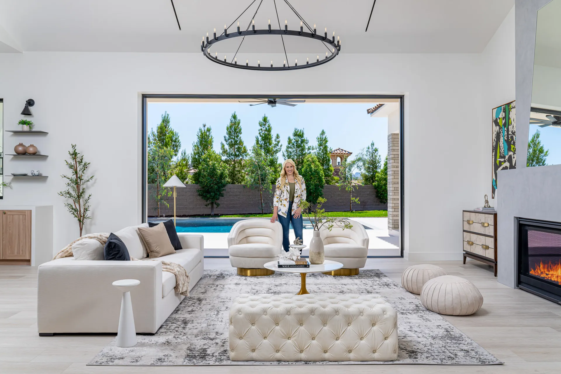 Modern living room with a white sofa, view of a pool and garden, and a woman standing in the room. The woman, Kelly Long, Lead Interior Designer, is wearing a green blouse and jeans. The room features a fireplace, a chandelier, and a rug, creating a stylish and comfortable space.