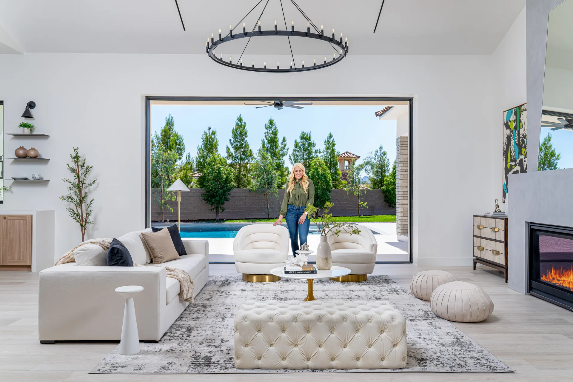 Modern living room with a white sofa, view of a pool and garden, and a woman standing in the room. The woman, Kelly Long, Lead Interior Designer, is wearing a green blouse and jeans. The room features a fireplace, a chandelier, and a rug, creating a stylish and comfortable space.