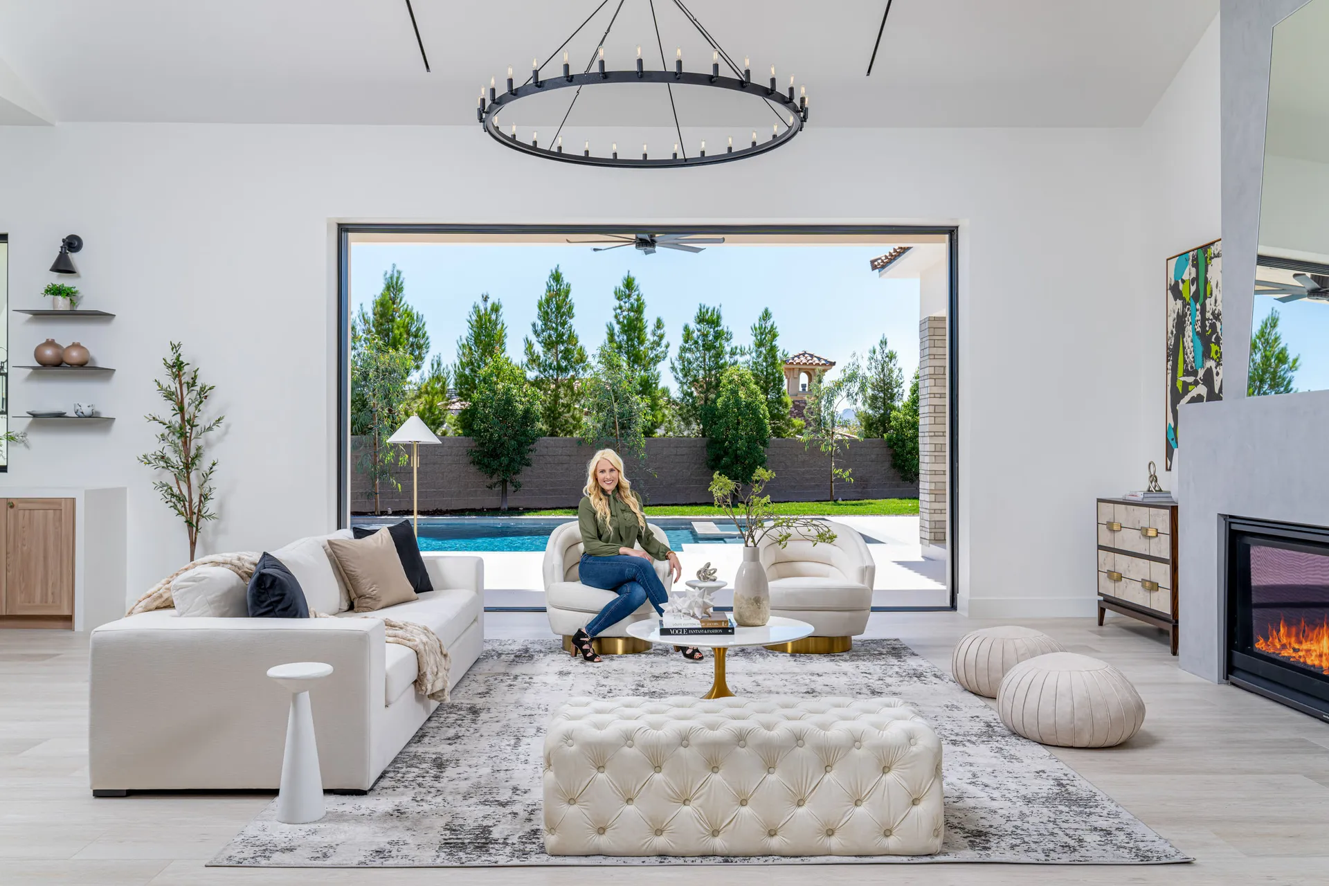 Modern living room with a white sofa, view of a pool and garden, and a woman sitting on the sofa. The woman, Kelly Long, Lead Interior Designer, is wearing a green blouse and jeans. The room features a fireplace, a chandelier, and a rug, creatinModern living room with a white sofa, view of a pool and garden, and a woman sitting on the sofa. The woman, Kelly Long, Lead Interior Designer, is wearing a green blouse and jeans. The room features a fireplace, a chandelier, and a rug, creating a stylish and comfortable space.g a stylish and comfortable space.