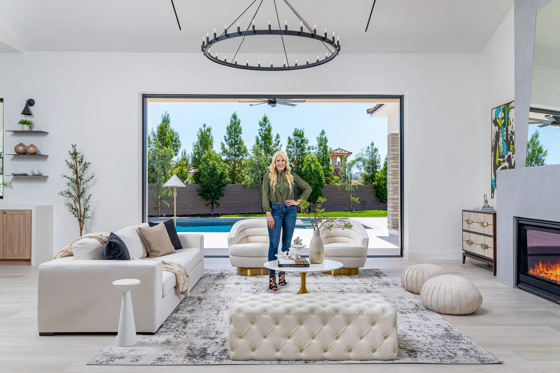 A blonde woman stands in a bright living area with a white sofa, modern chairs, and large sliding doors leading to a backyard pool. A ring-shaped chandelier hangs overhead.