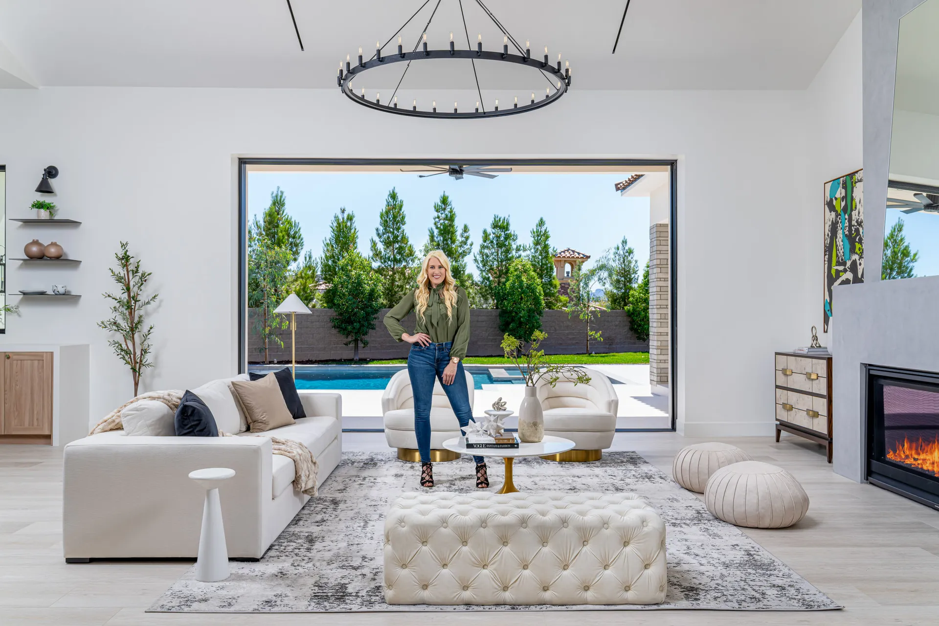 Modern living room with a white sofa, view of a pool and garden, and a woman standing in the room. The woman, Kelly Long, Lead Interior Designer, is wearing a green blouse and jeans. The room features a fireplace, a chandelier, and a rug, creating a stylish and comfortable space.