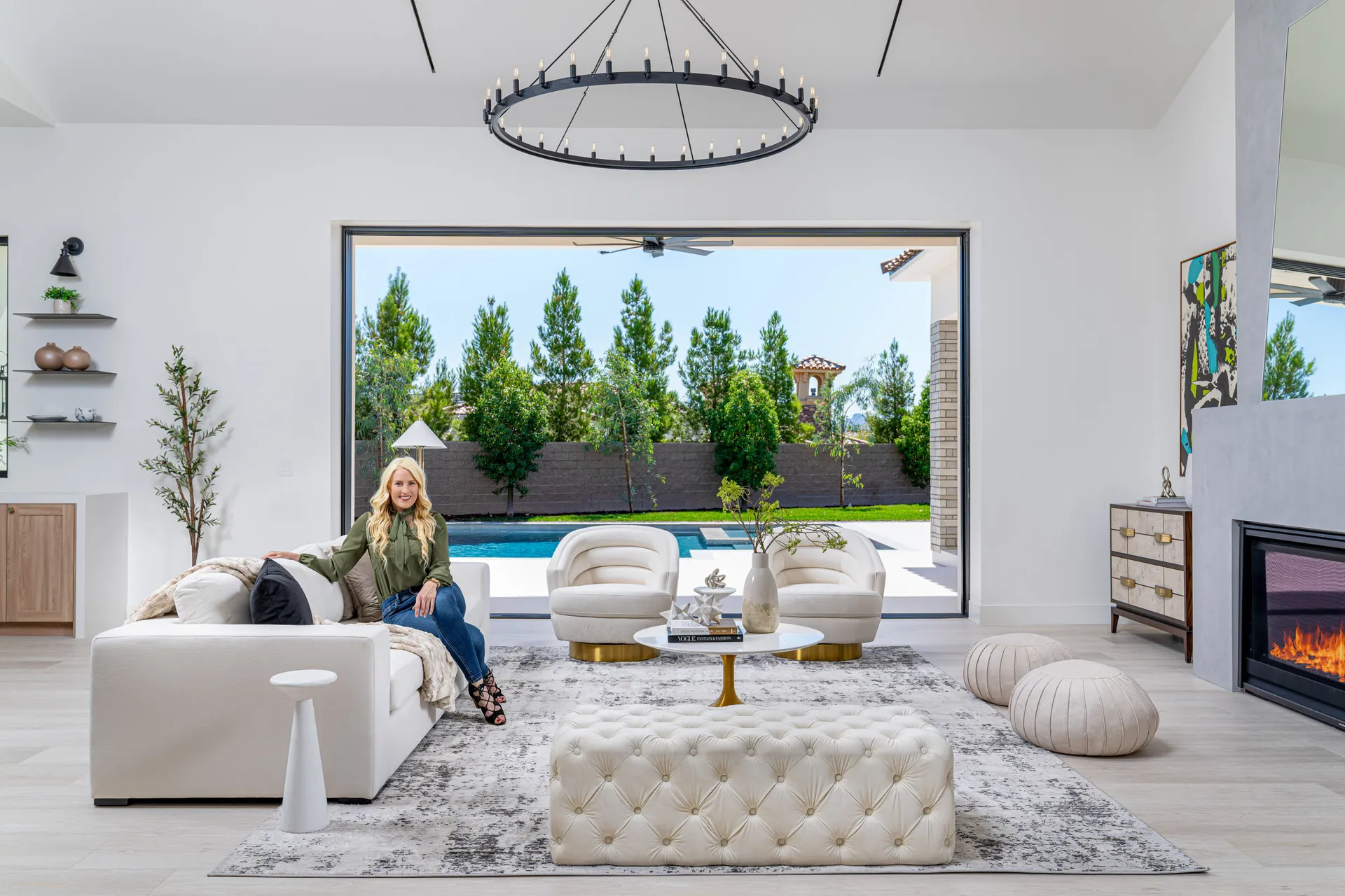 Modern living room with a white sofa, view of a pool and garden, and a woman sitting on the sofa. The woman, Kelly Long, Lead Interior Designer, is wearing a green blouse and jeans. The room features a fireplace, a chandelier, and a rug, creating a stylish and comfortable space.