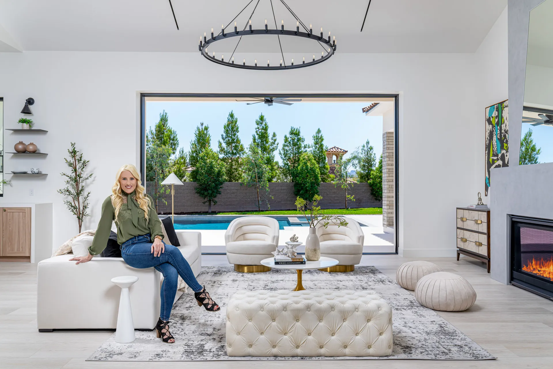 A blonde woman sits on a white sofa in a bright living area. Large sliding doors open to a backyard with a swimming pool, and a circular chandelier hangs overhead.