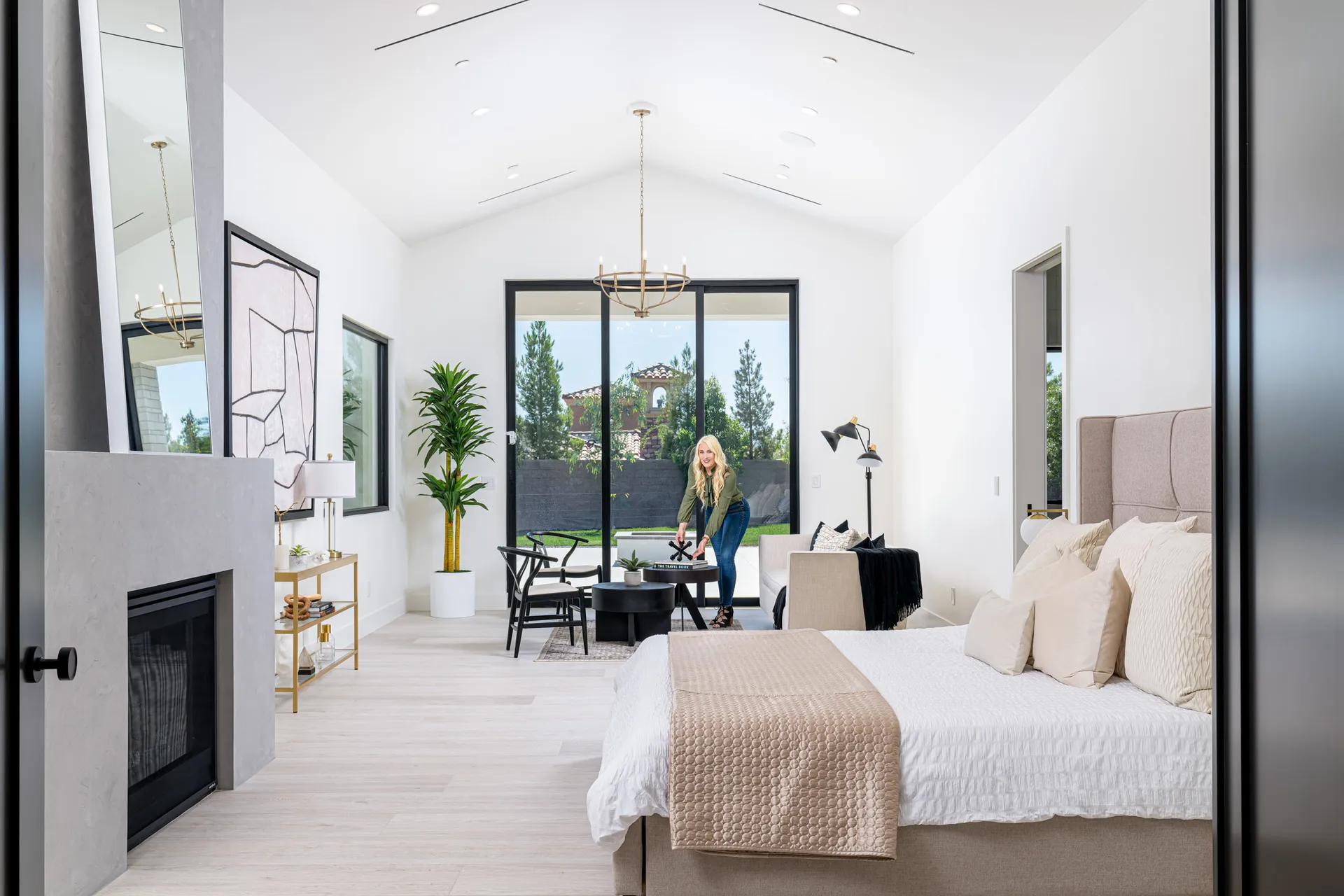 A bright bedroom with a vaulted ceiling, a fireplace, and a bed dressed in neutral bedding. A blonde woman arranges decor on a small round table near sliding glass doors.