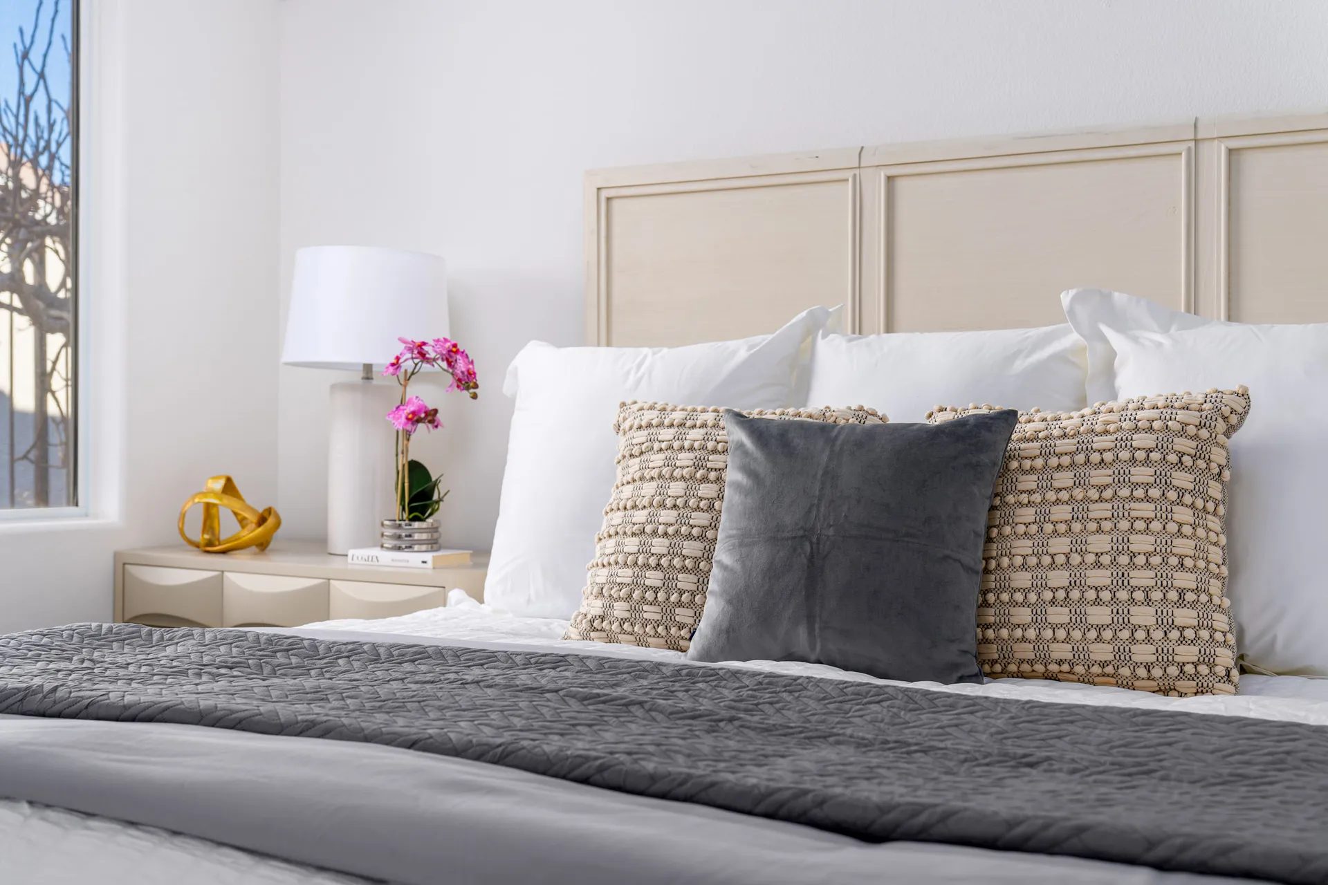 A stylish bedroom with a neutral wood headboard, layered bedding in soft grays and whites, and a nightstand adorned with fresh orchids and decor.