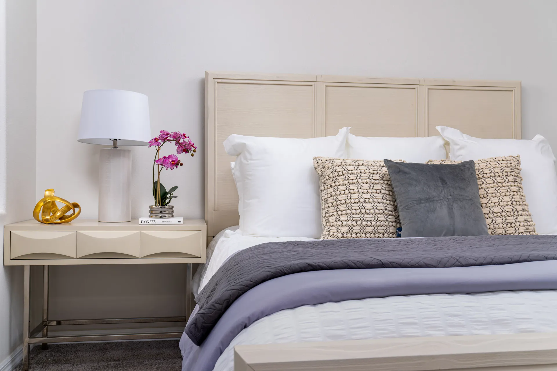A modern bedroom with a neutral wood headboard, plush bedding, a stylish nightstand, and decorative accents including a white lamp and fresh orchids.