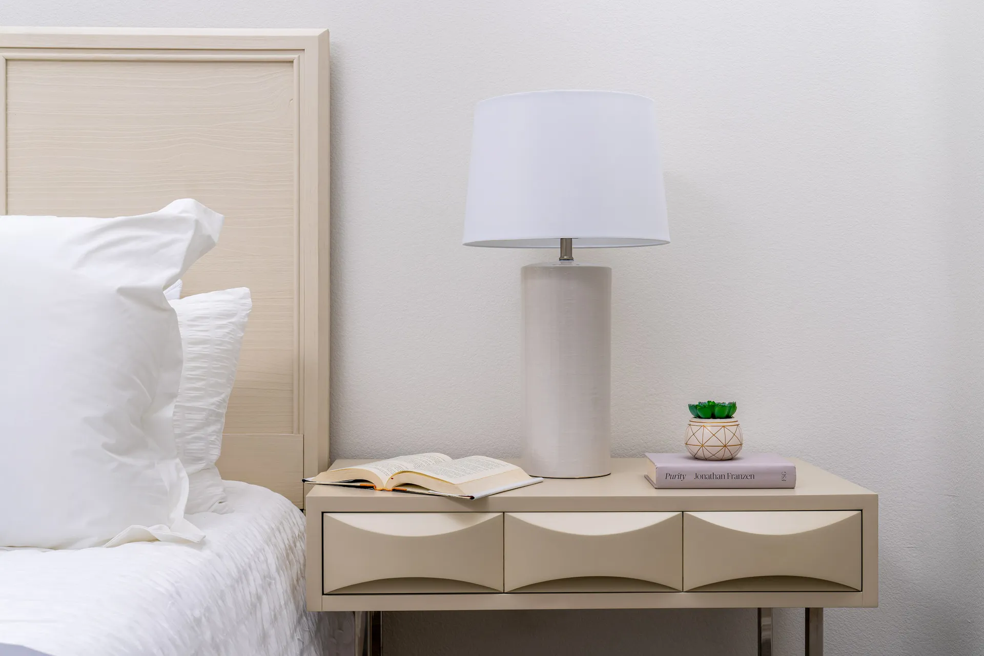 A modern bedroom nightstand with a white lamp, an open book, a small potted plant, and a neatly made bed with crisp white linens.