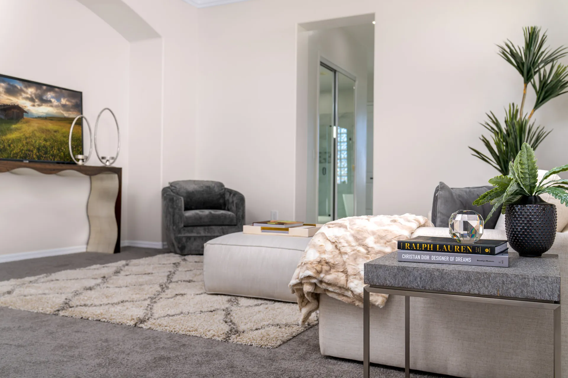 A cozy bedroom lounge area with a plush ottoman, soft throw blanket, modern decor, and a sculptural console table beneath a mounted TV.