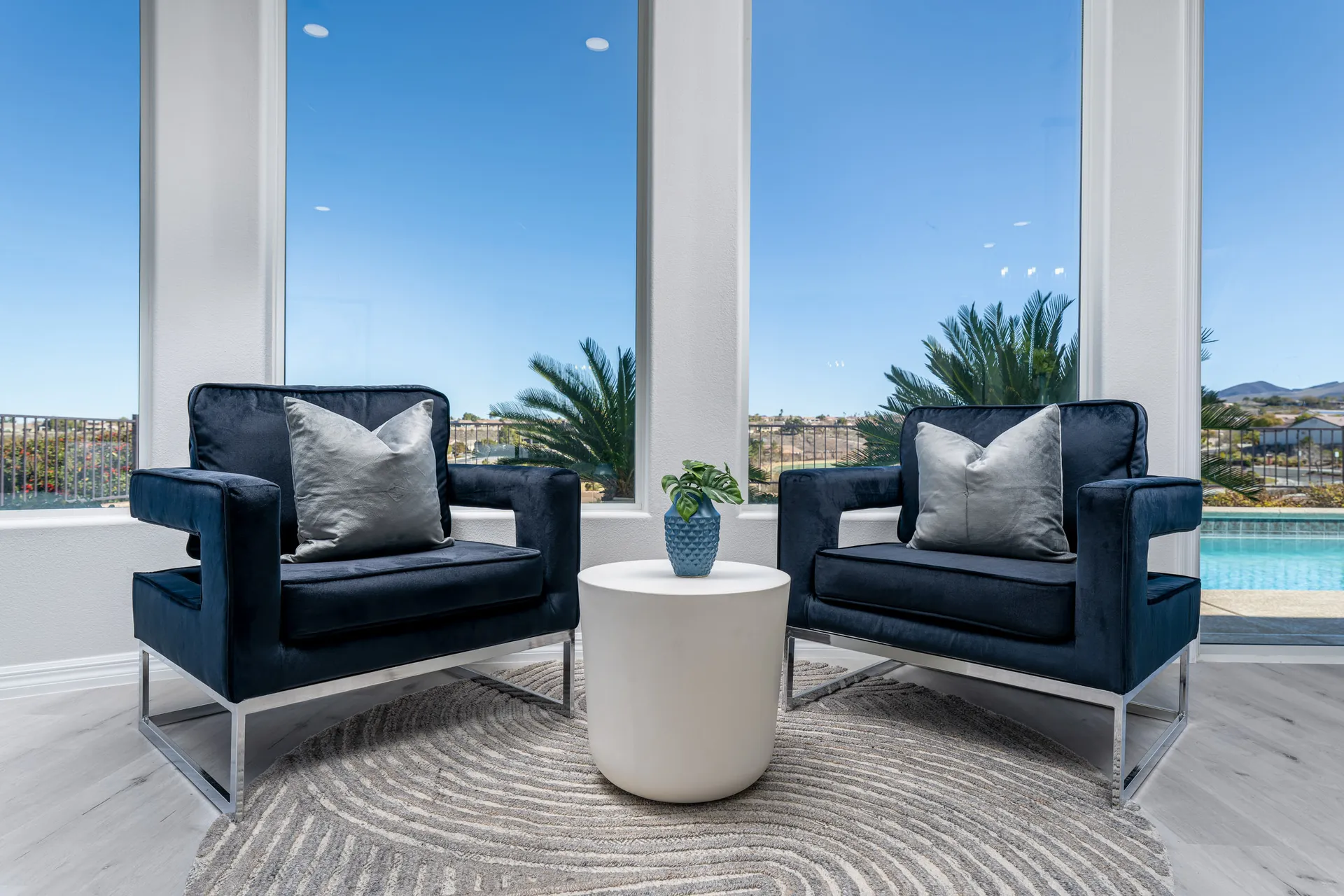 A cozy seating nook with two navy velvet chairs, a white round side table, and floor-to-ceiling windows overlooking a pool and palm trees.
