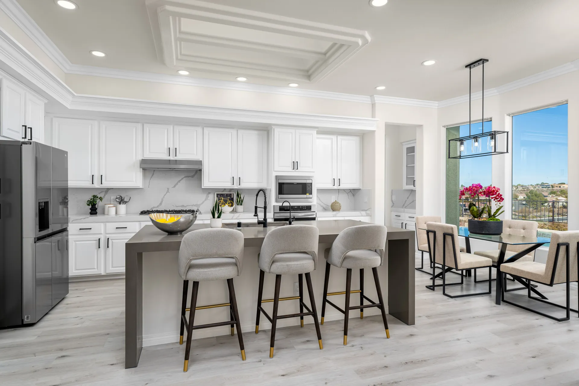 A modern kitchen with a large island, upholstered barstools, white cabinetry, and a bright dining area with large windows overlooking the outdoors.