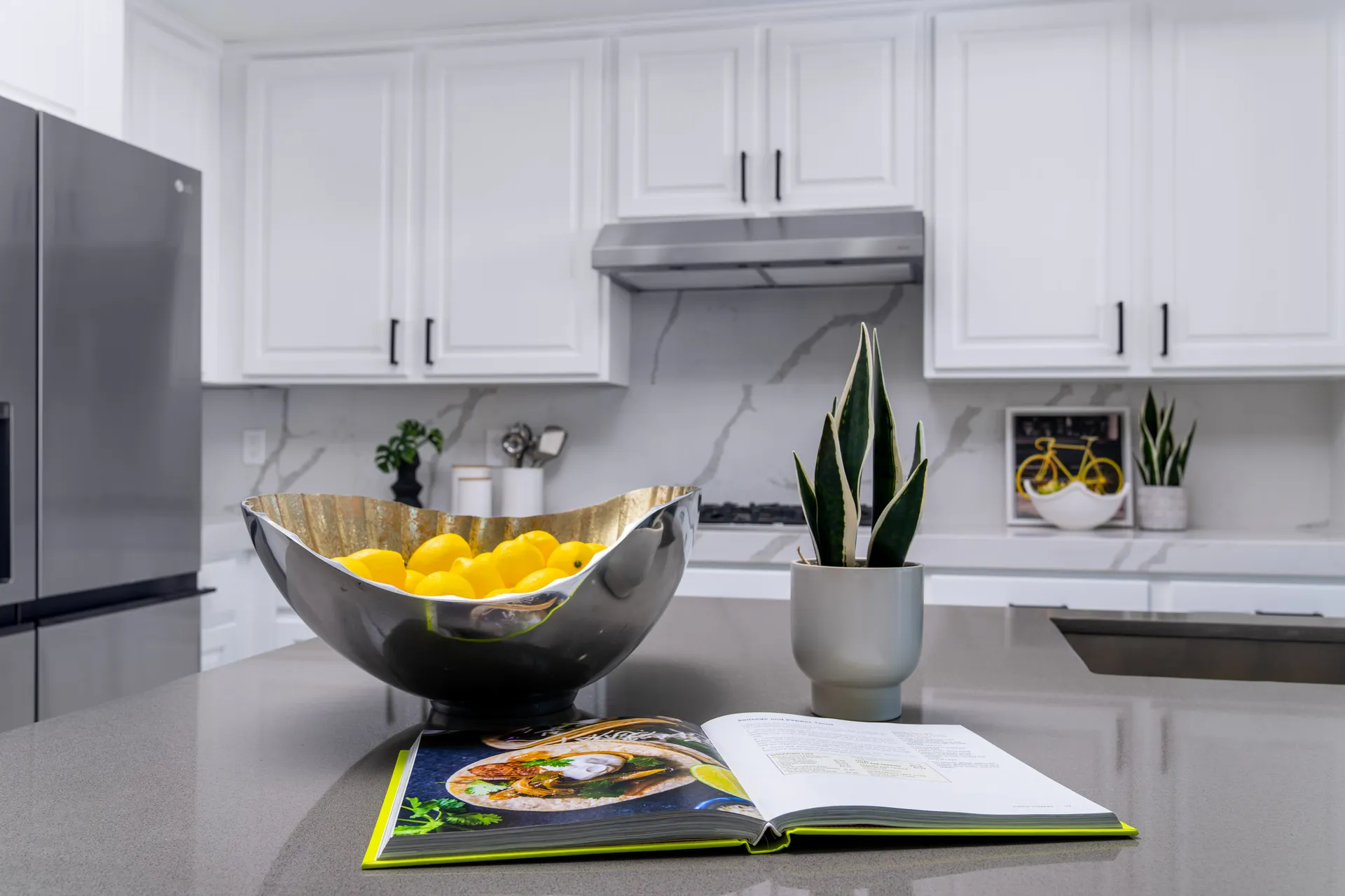 A modern kitchen with white cabinetry, marble backsplash, a dark quartz island, and stylish decor featuring lemons, a plant, and an open cookbook.