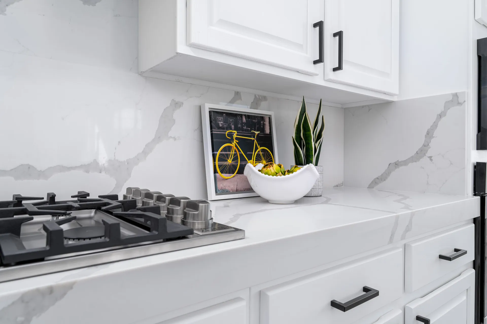 A sleek white kitchen with marble countertops, black hardware, and a stylish countertop display featuring a framed bicycle print and greenery.